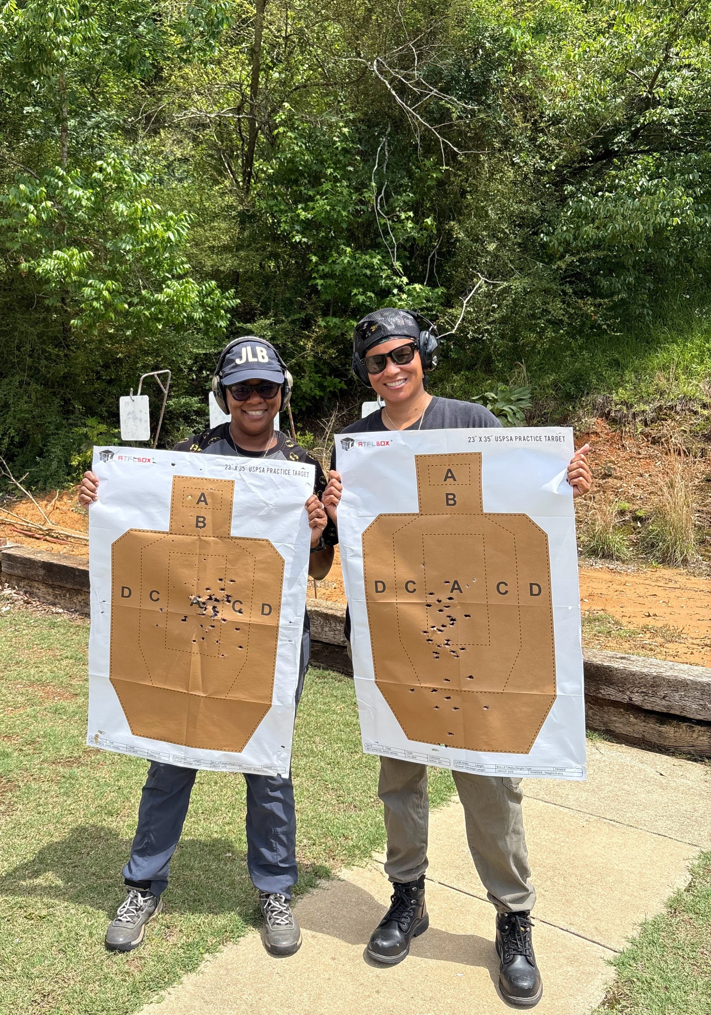 Two women smiling outdoors at a shooting range, holding paper target sheets with bullet holes, wearing protective ear gear and sunglasses, with greenery and trees in the background.