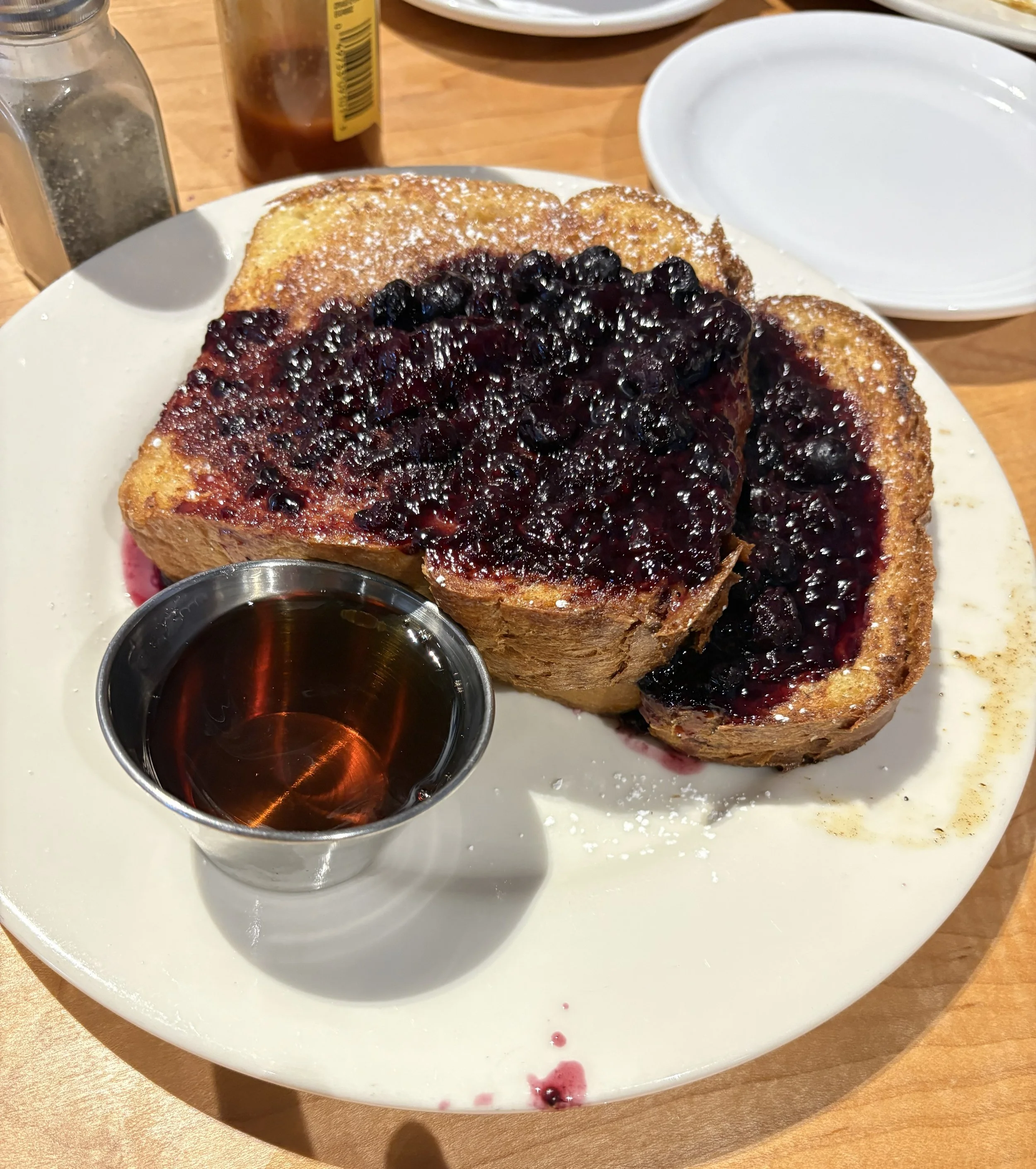 A plate with a slice of French toast topped with blueberry compote, a small metal cup of syrup, and a white bowl with empty space. In the background, there are salt and pepper shakers, a sauce bottle, and dishes on a wooden table.