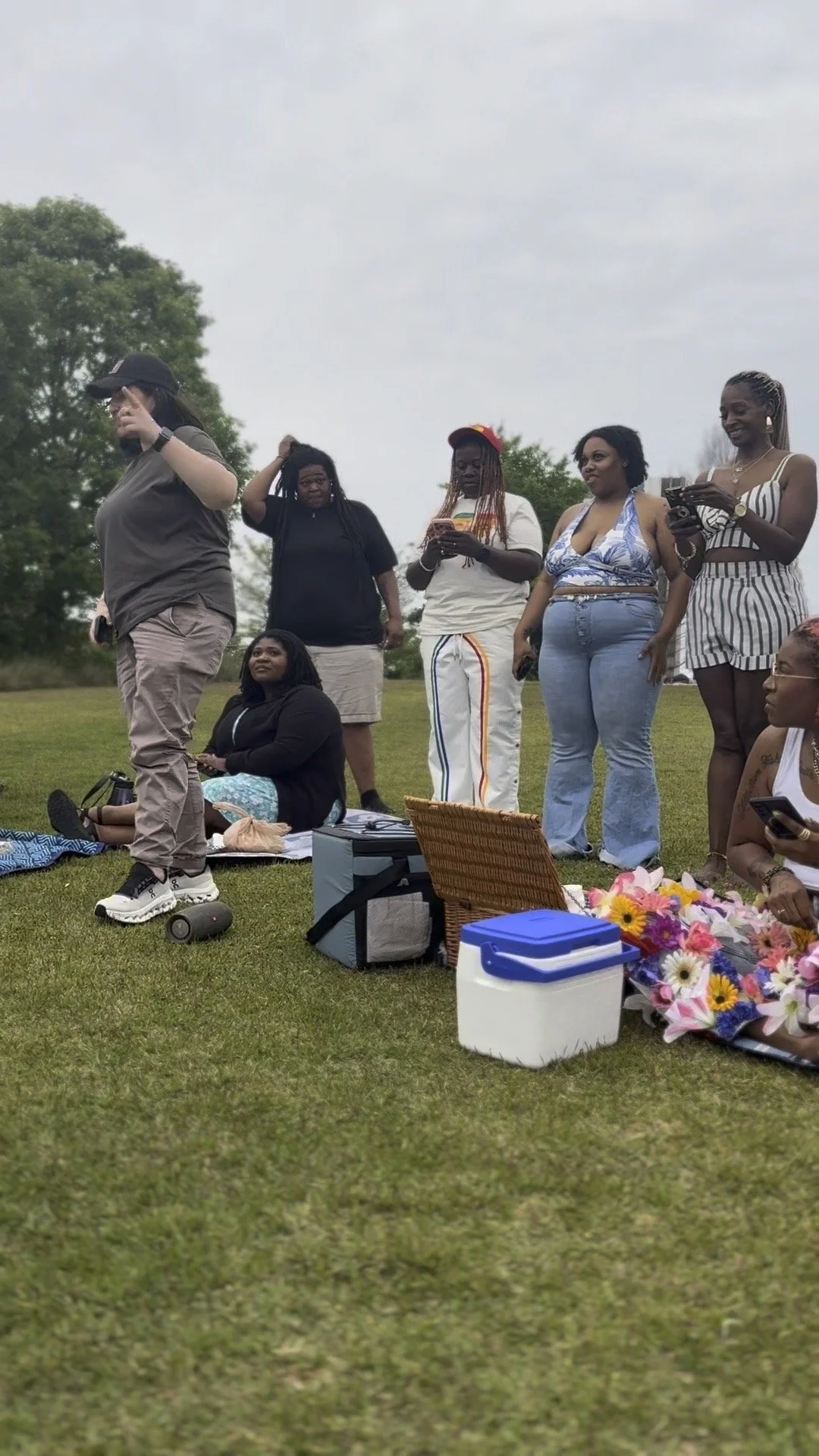 Group of women gathered outdoors on a grassy field, some standing and some sitting, with assorted bags and flowers nearby.
