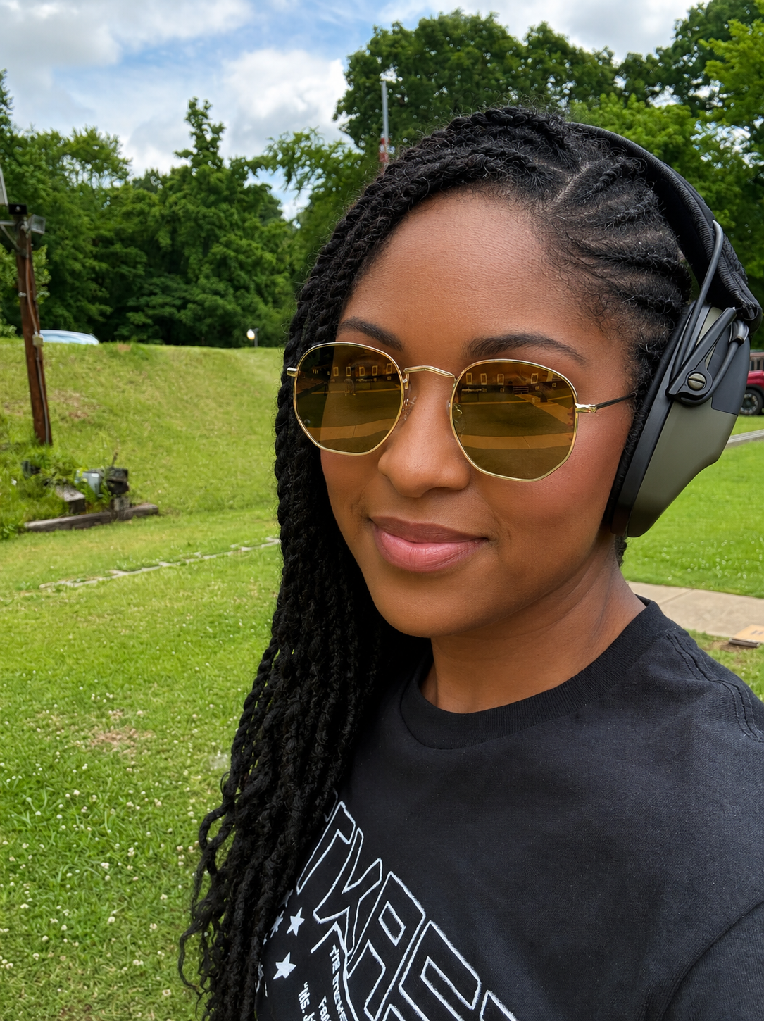A woman with braided hair and sunglasses wearing noise-canceling headphones outdoors on a grassy area with trees and a cloudy sky in the background.