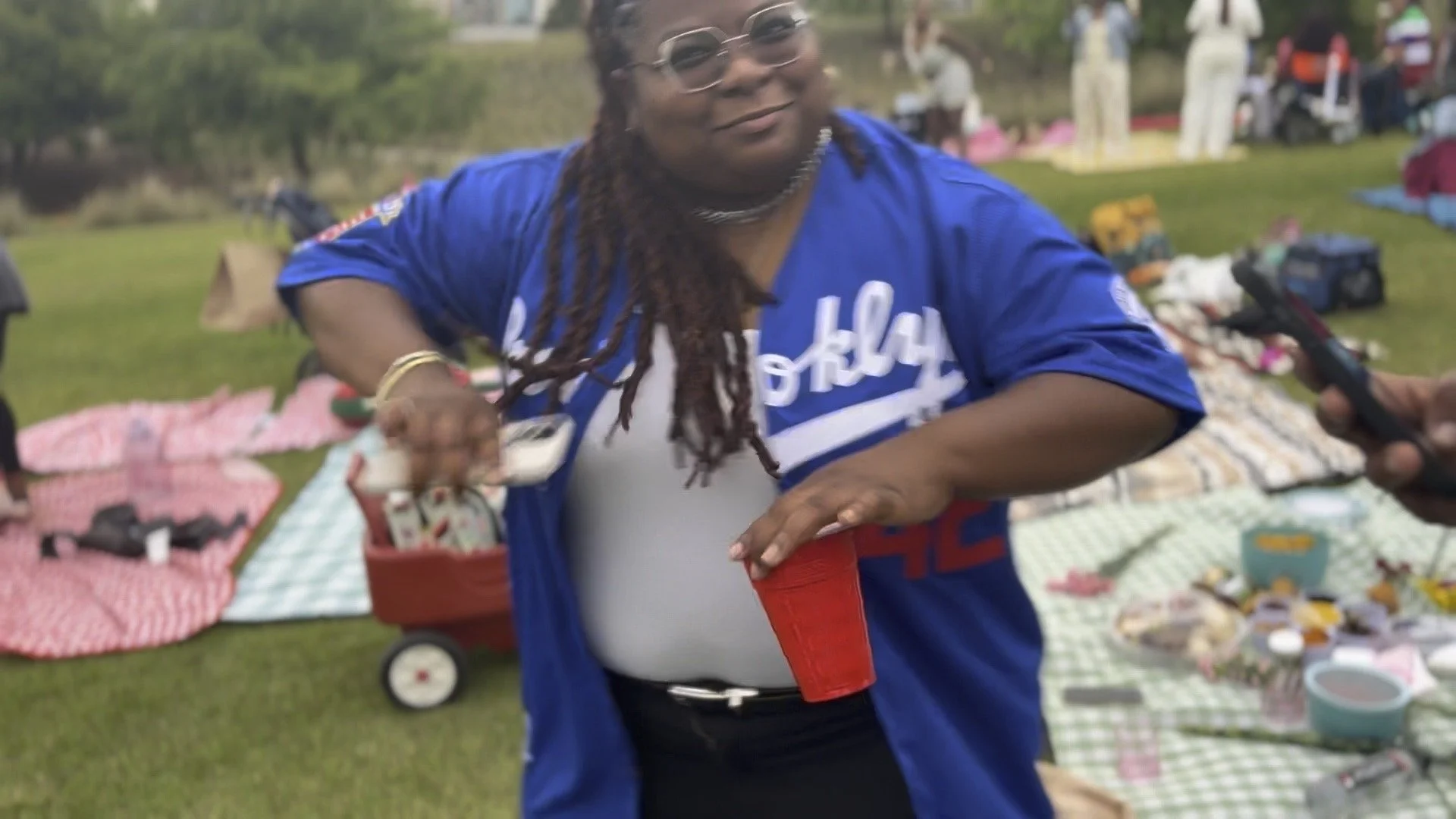 A woman with long dreadlocks, glasses, and a blue sports jersey pouring a drink into a red cup at an outdoor gathering with blankets, picnic items, and other people in the background.