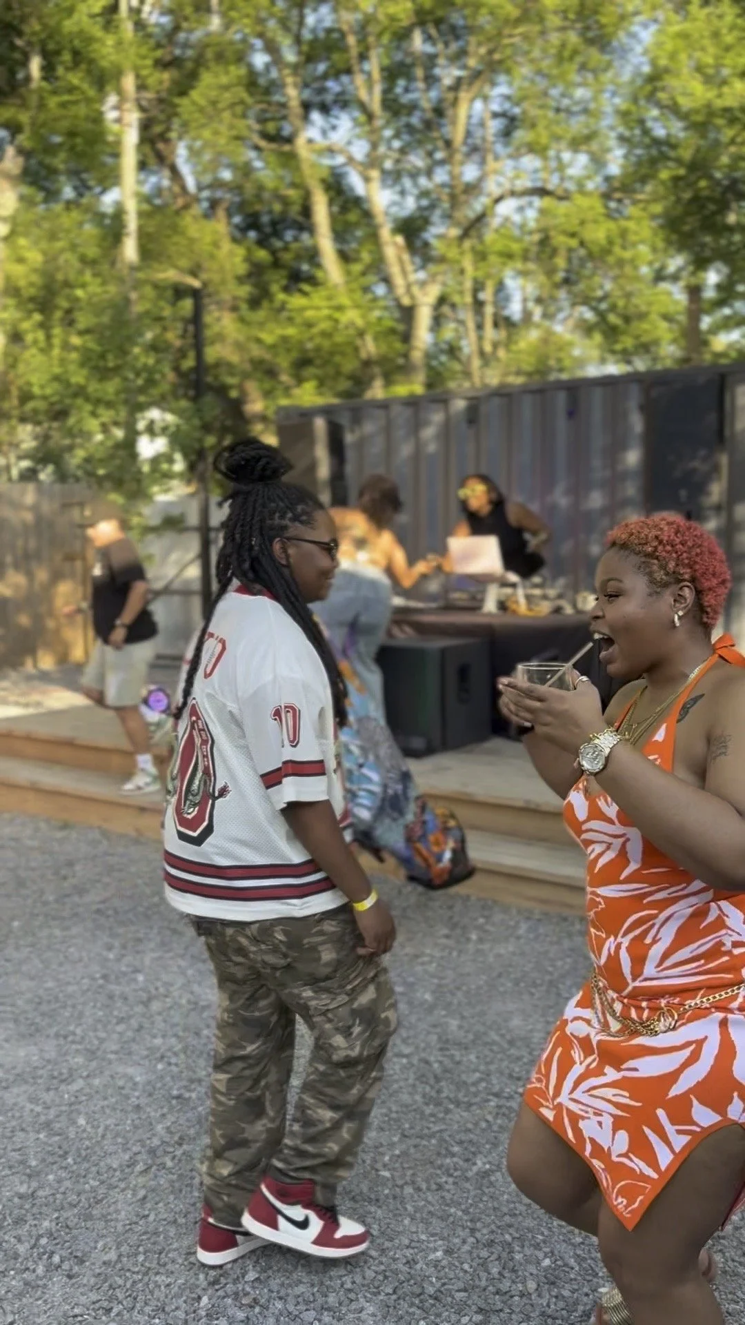Two women are talking and laughing outdoors at a social gathering, with a DJ playing music in the background and people in casual summer clothing.