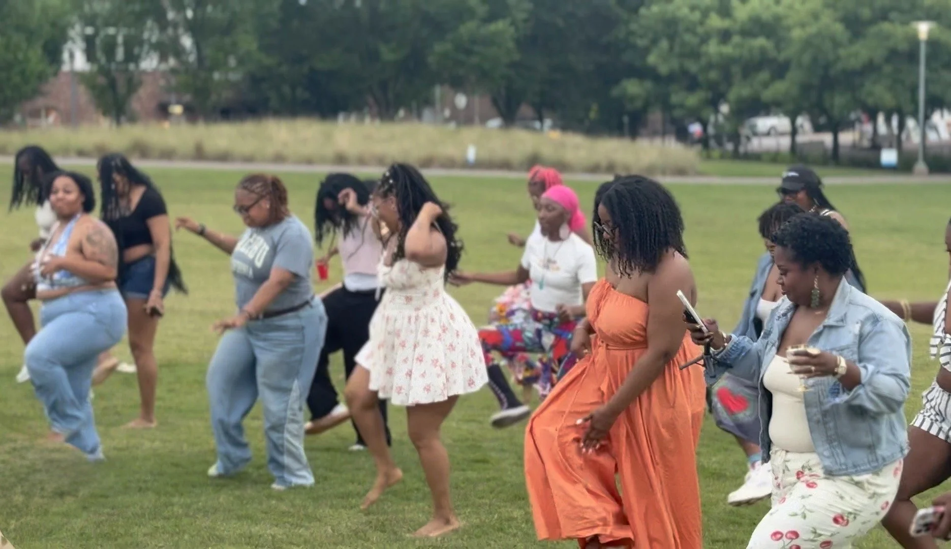 A group of women dancing outdoors on a grassy field during a sunny day, some are looking at their phones, others are smiling and enjoying the moment, with trees and a park in the background.