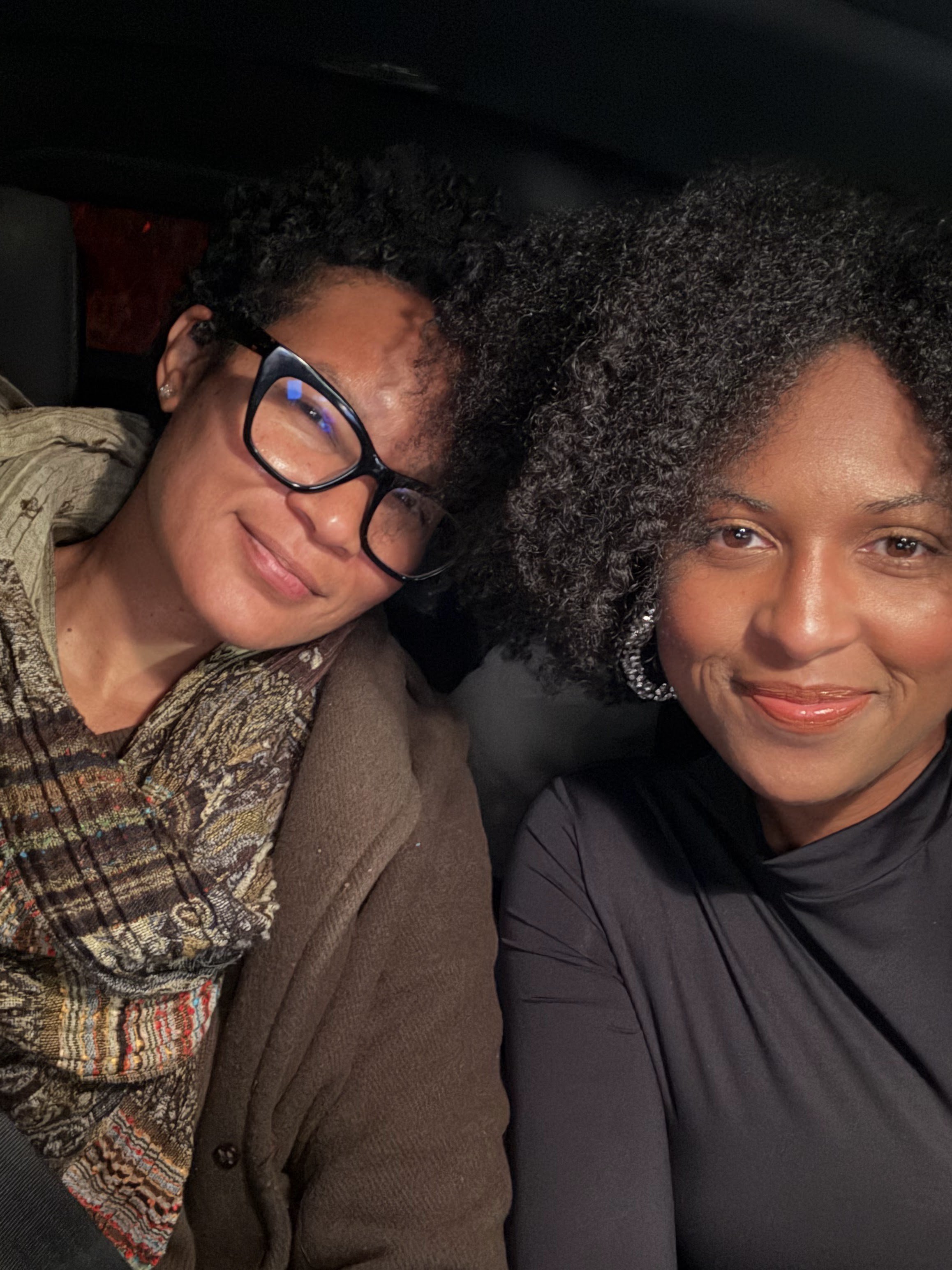 Close-up of two women sitting side by side, smiling, in a dark setting. Both women have curly hair, and the woman on the left is wearing glasses and earrings. The woman on the right has hoop earrings and is wearing a black top.