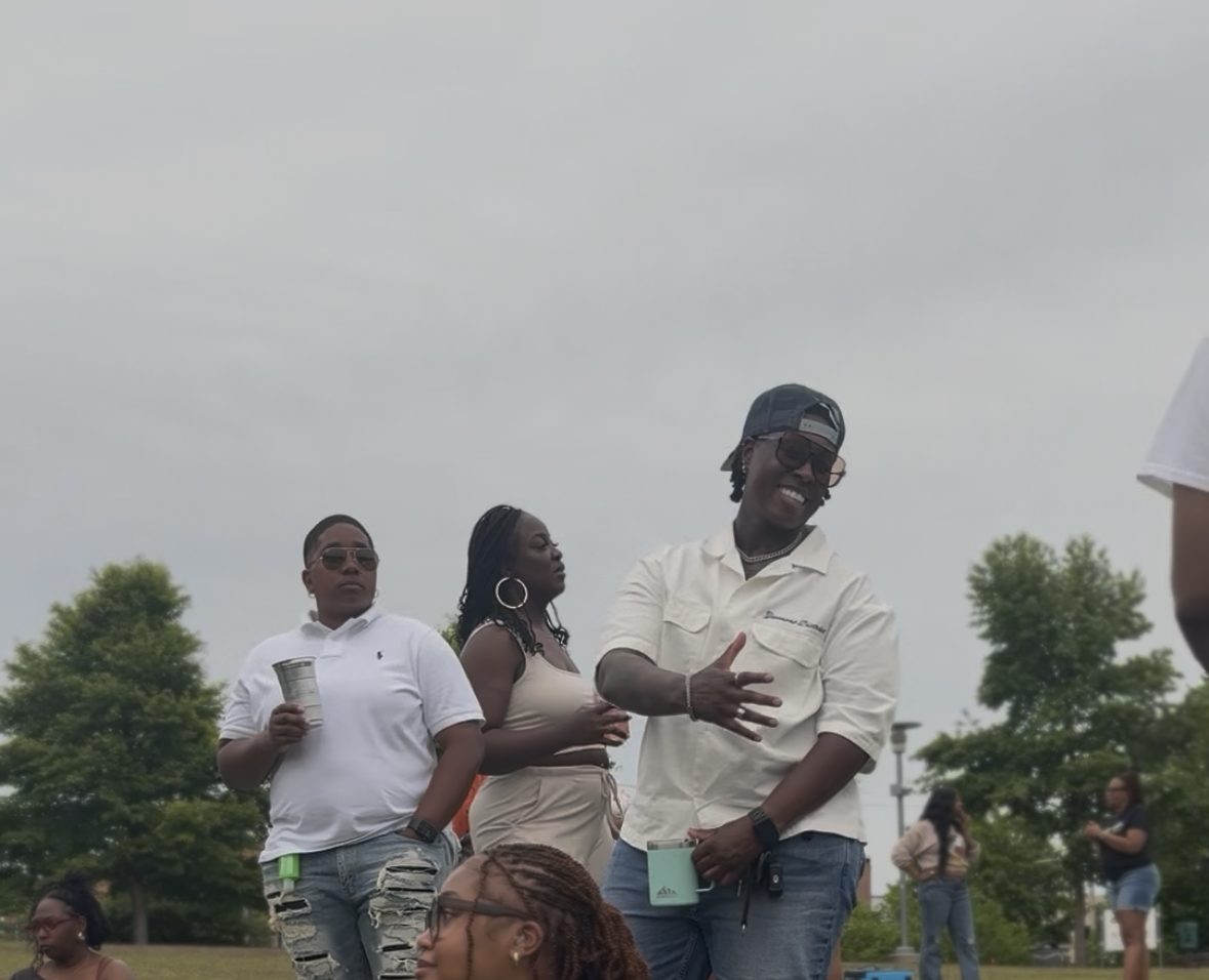 Group of people outdoors, one smiling man in a white shirt and cap speaking, others standing and holding drinks, trees and cloudy sky in background.