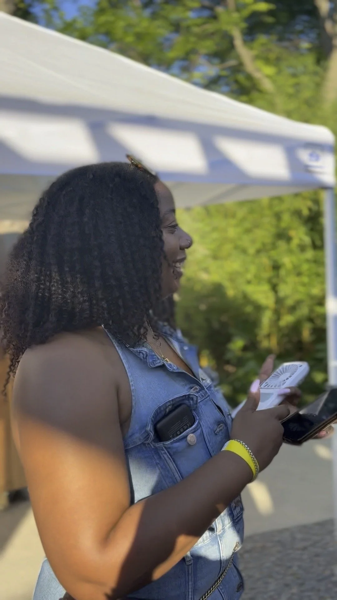 A woman with curly hair wearing a sleeveless denim vest and holding a phone, smiling outdoors under a white canopy.