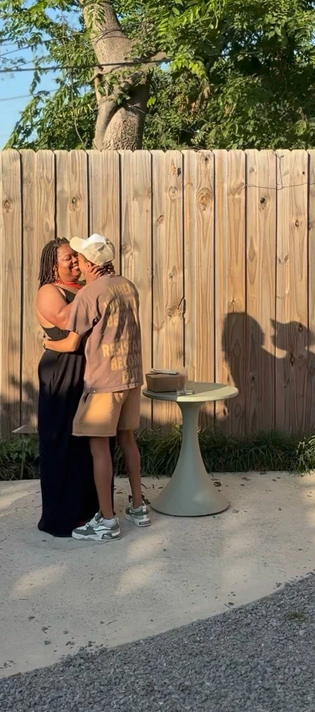A couple hugs and kisses outdoors in front of a wooden fence and a large tree. They are standing on a concrete surface next to a small round table with some boxes on it. Their shadows are visible on the fence.