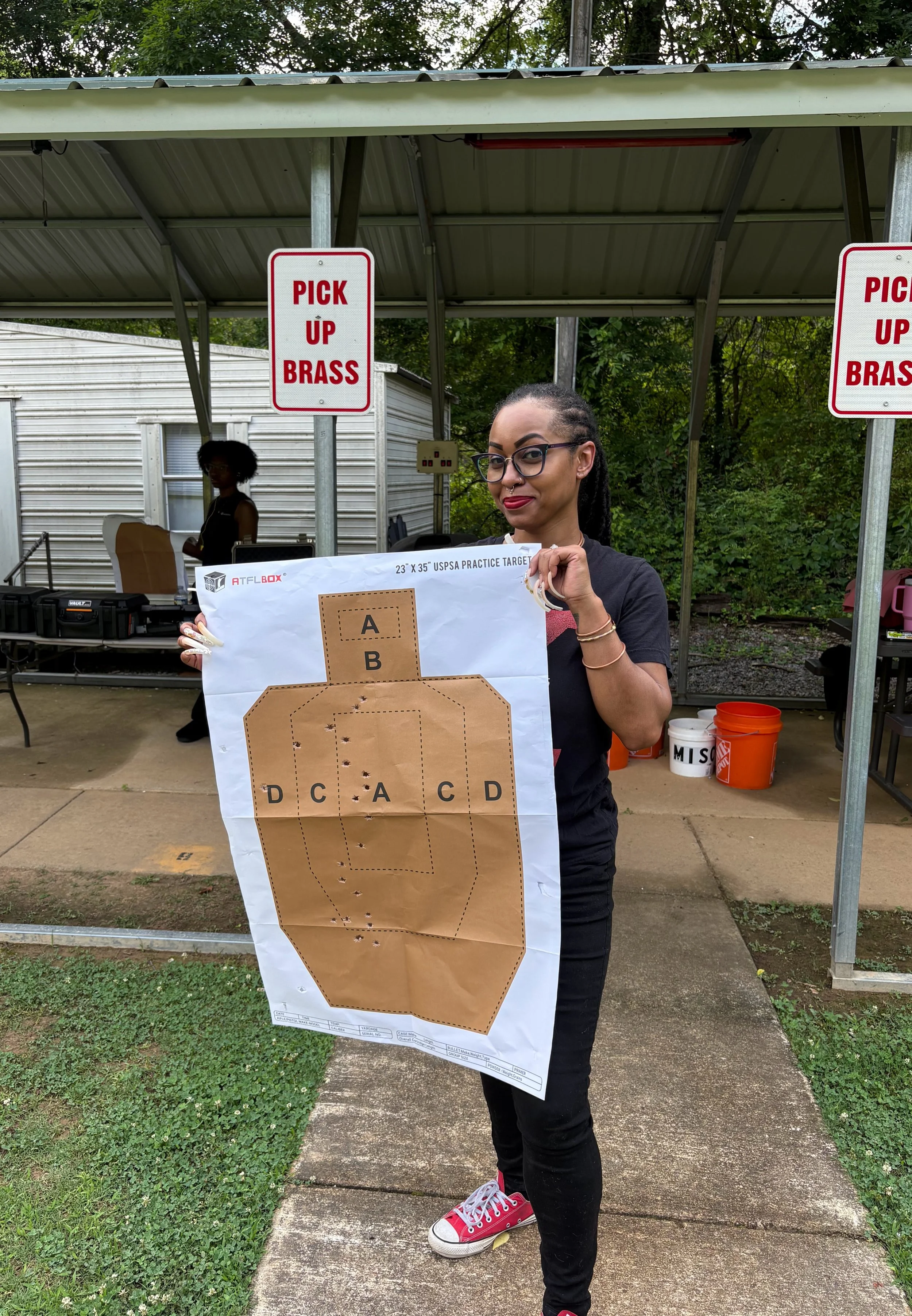 A woman with glasses and red shoes holding a large paper target outdoors with a metal shelter and trees in the background.