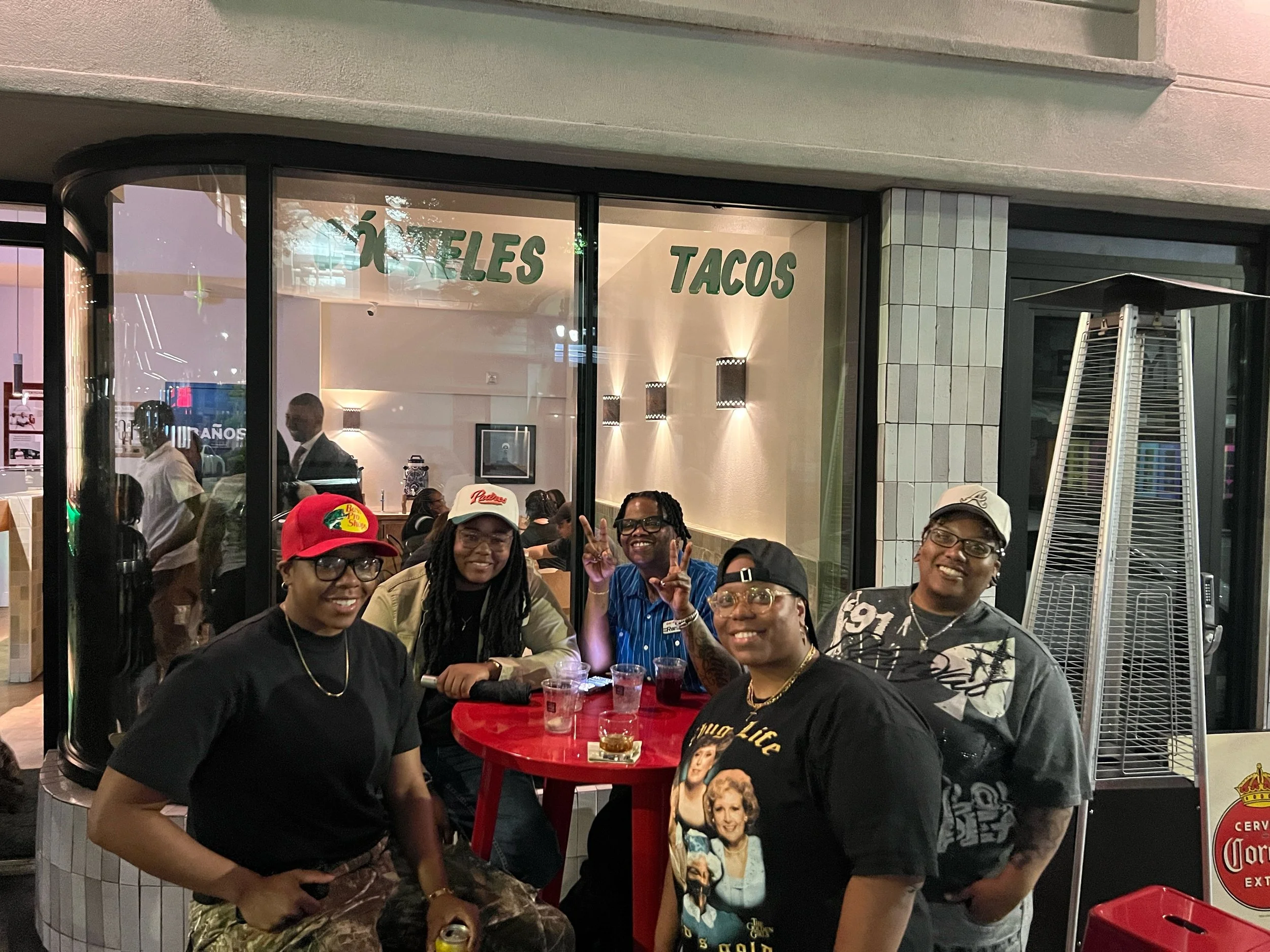 Group of people smiling at a table inside a restaurant with glass windows and sign that reads 'TACOS' and 'WHOLESALE'.