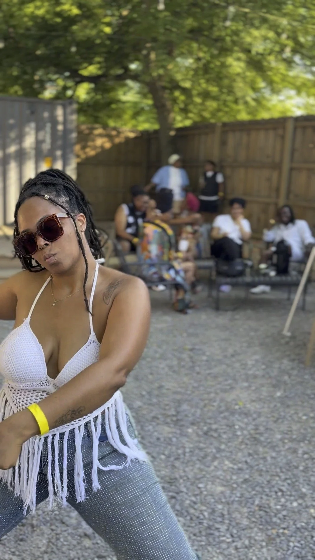 Person in white crochet top and sunglasses in front with a group of people sitting and standing in backyard with trees and wooden fence