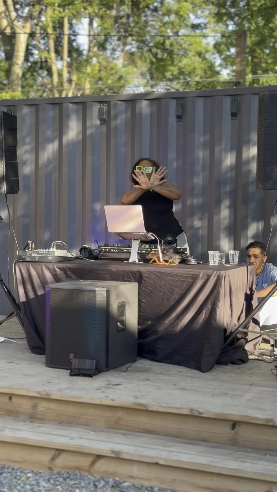Woman DJ wearing sunglasses, standing behind a DJ setup with a laptop and electronic equipment, outdoors during daytime, with man sitting at the side, stage with wooden floor, large speakers, and trees in the background.