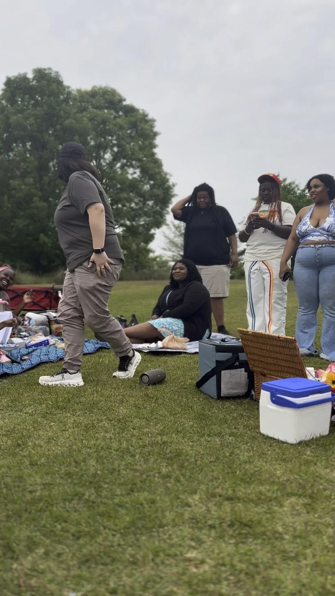 A group of women having a picnic outdoors on a grassy field with trees in the background. One woman is standing and speaking, while others are sitting or standing, some smiling and others looking at their phones.