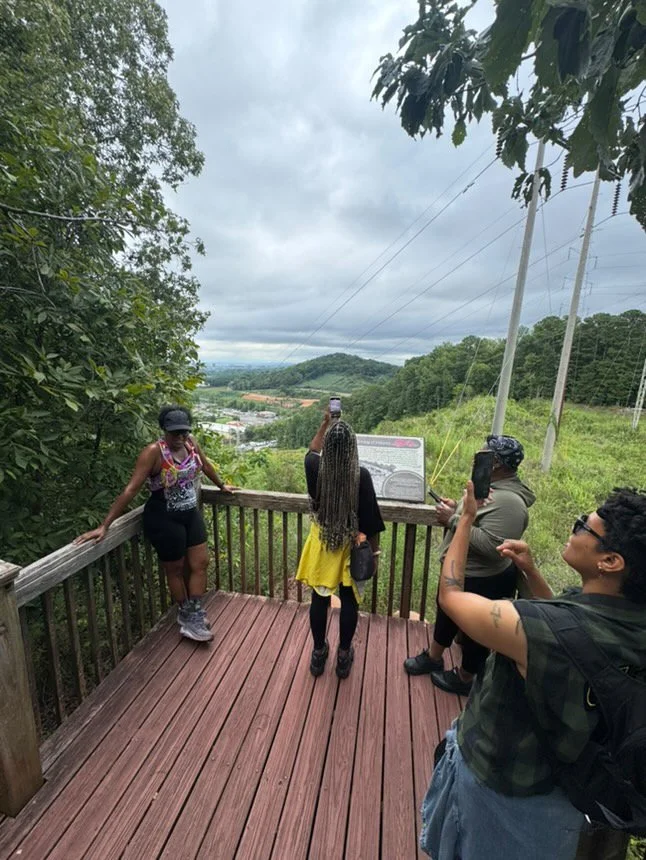 Four people at a scenic lookout on a wooden deck, overlooking mountainous terrain and cloudy sky, with trees and power lines in the background.
