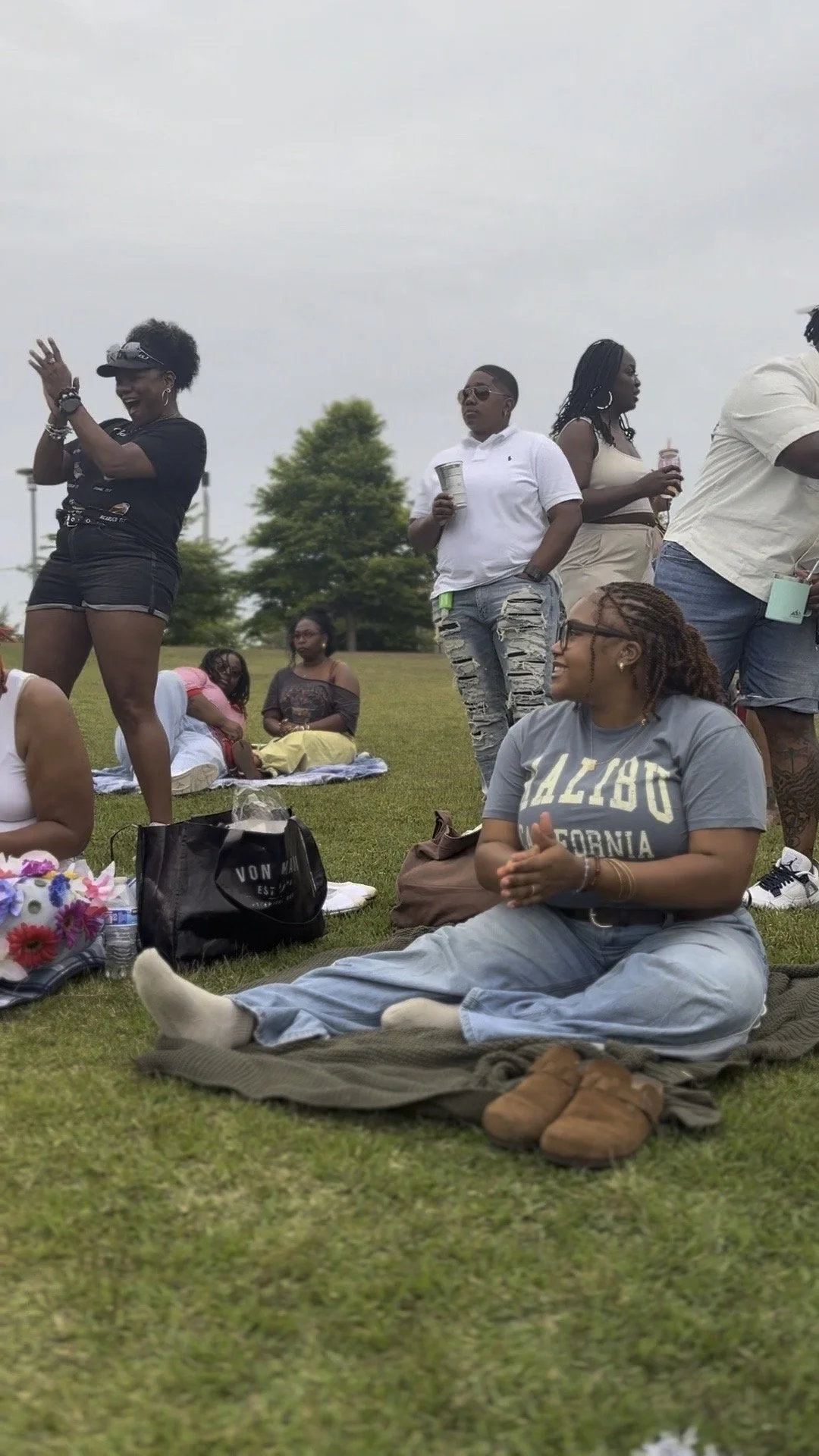 Group of people gathered outdoors on the grass, some sitting and some standing, enjoying a social event or picnic on a cloudy day.