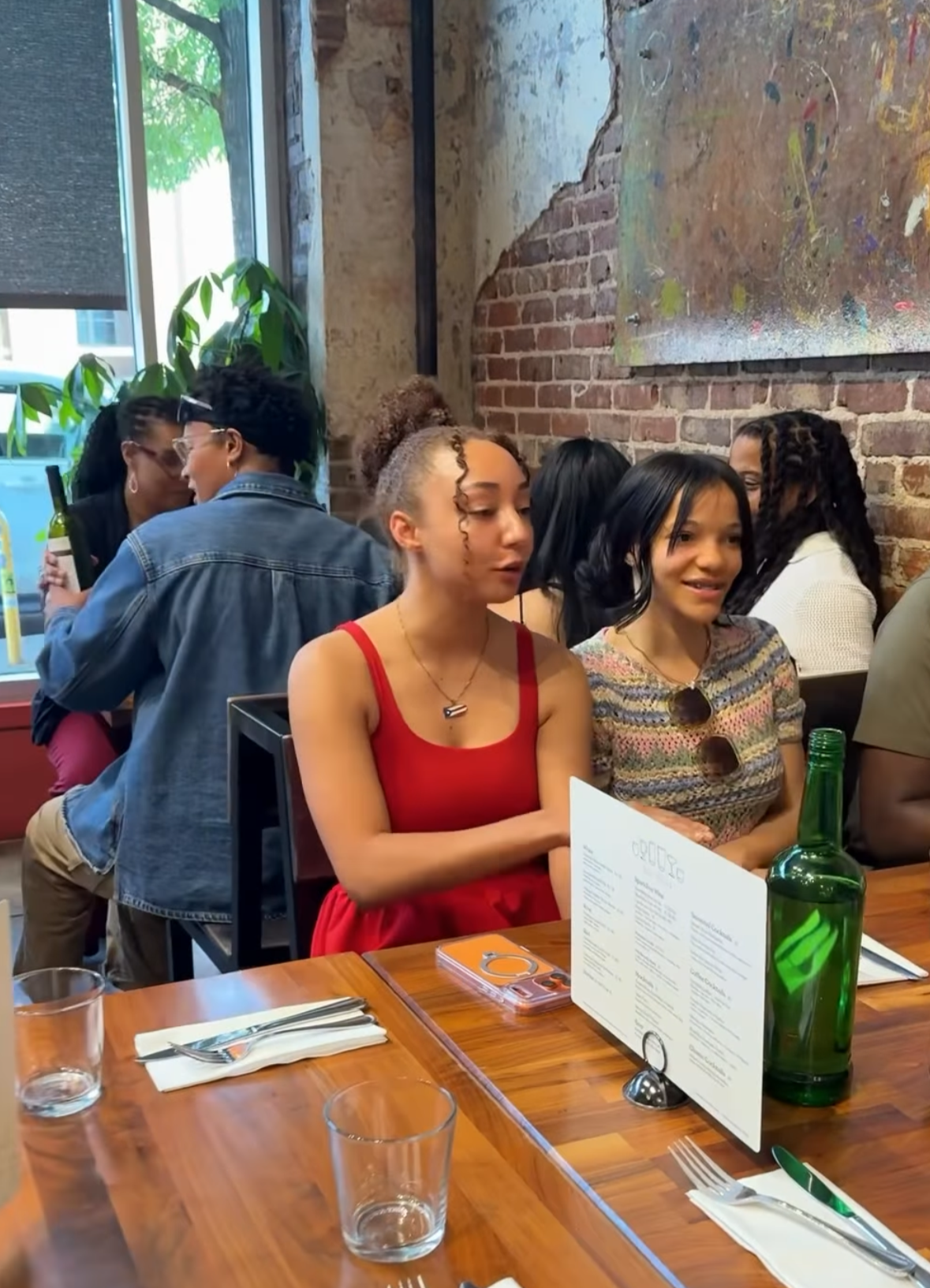 Four women sitting at a restaurant table, engaged in conversation. Two women in the foreground, one in a red dress, and the other in a patterned top. Two women in the background near the window, one holding a bottle of wine. The restaurant has expose