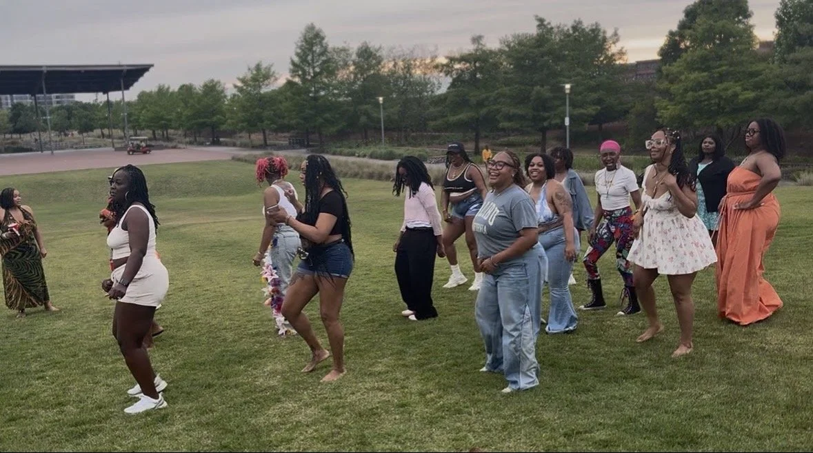 Group of women dancing on a grassy field during an outdoor event, with trees and a pavilion visible in the background.