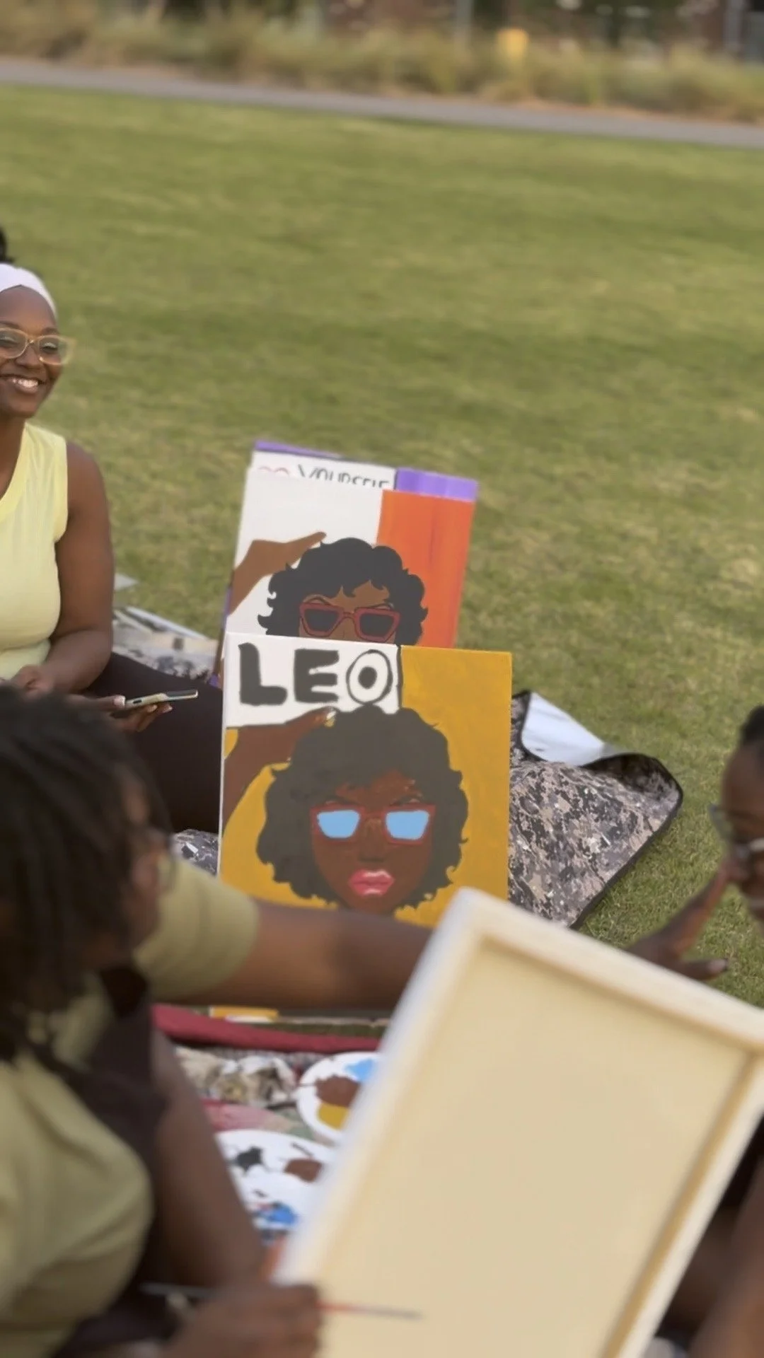 Group of people outdoor, sitting on blankets, with colorful artwork featuring women with curly hair and glasses, labeled 'LEO' and another with clothing and accessories, on display.