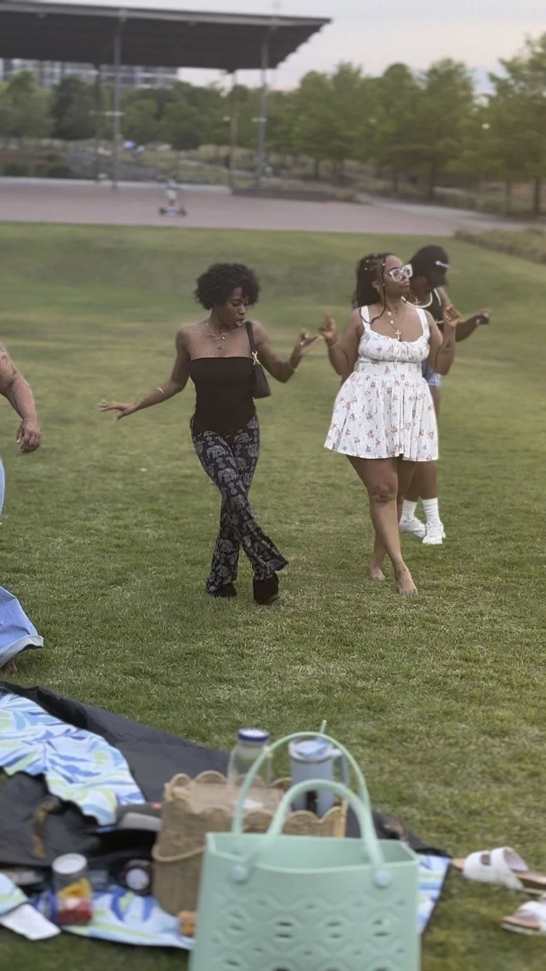 Three women dancing outdoors on a grassy area during a gathering, with picnic items and bags in the foreground.