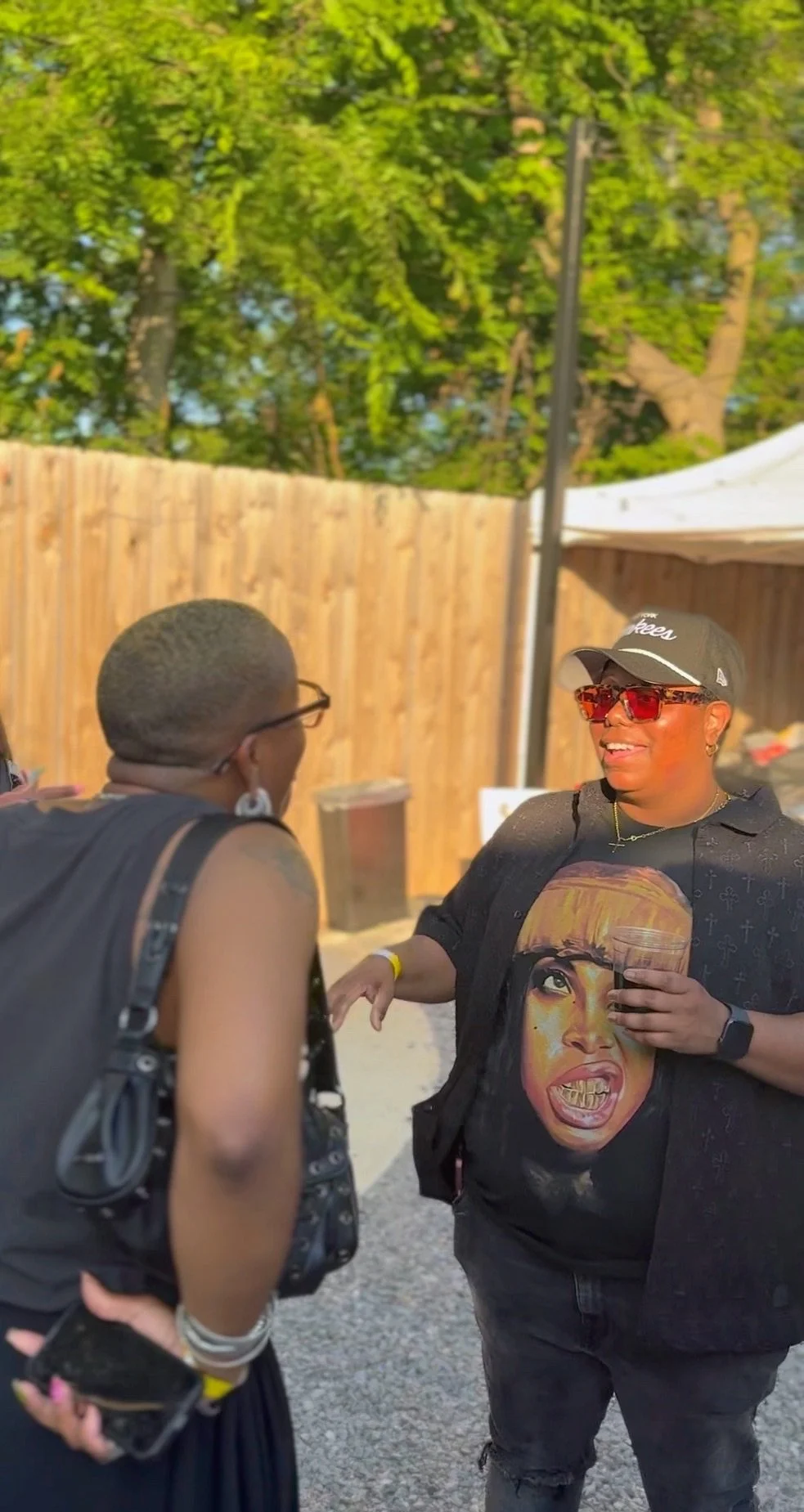Two women engaged in conversation outdoors near a wooden fence, with trees in the background; one woman is wearing a black cap, red sunglasses, and a black shirt with a comic-style face design, holding a drink; the other woman has short hair, glasses