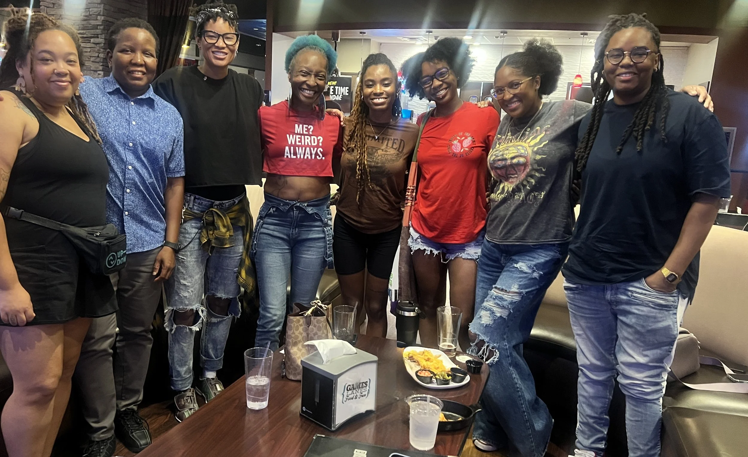 Group of women standing together at a social gathering in a restaurant, smiling and posing for the photo, with food and drinks on the table in front of them.