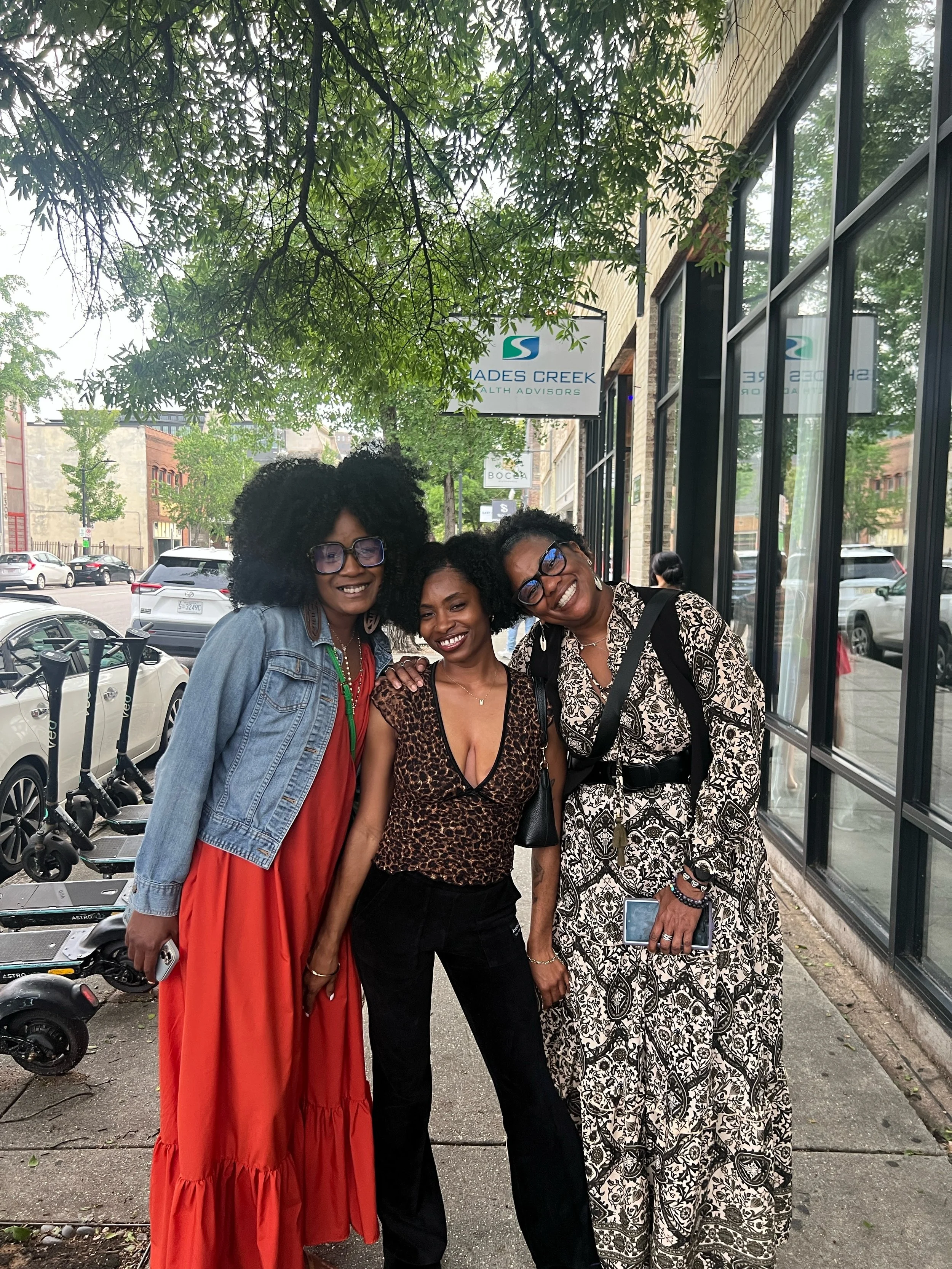 Three women standing outside on a sidewalk, smiling, with cars, electric scooters, trees, and a building with large windows behind them.