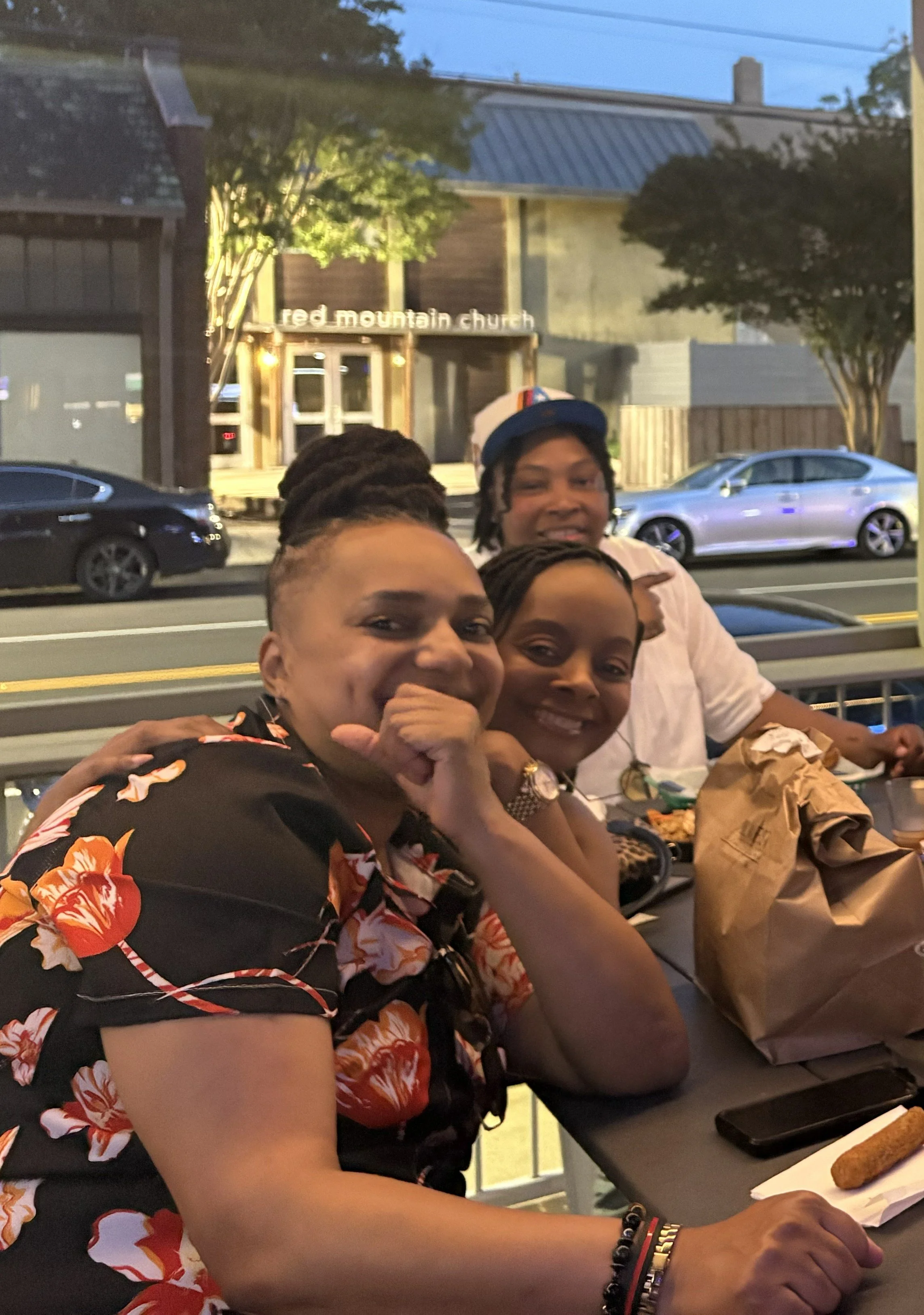 Four smiling people sitting together at a table outside in front of the Red Mountain Church. The setting appears to be during early evening.