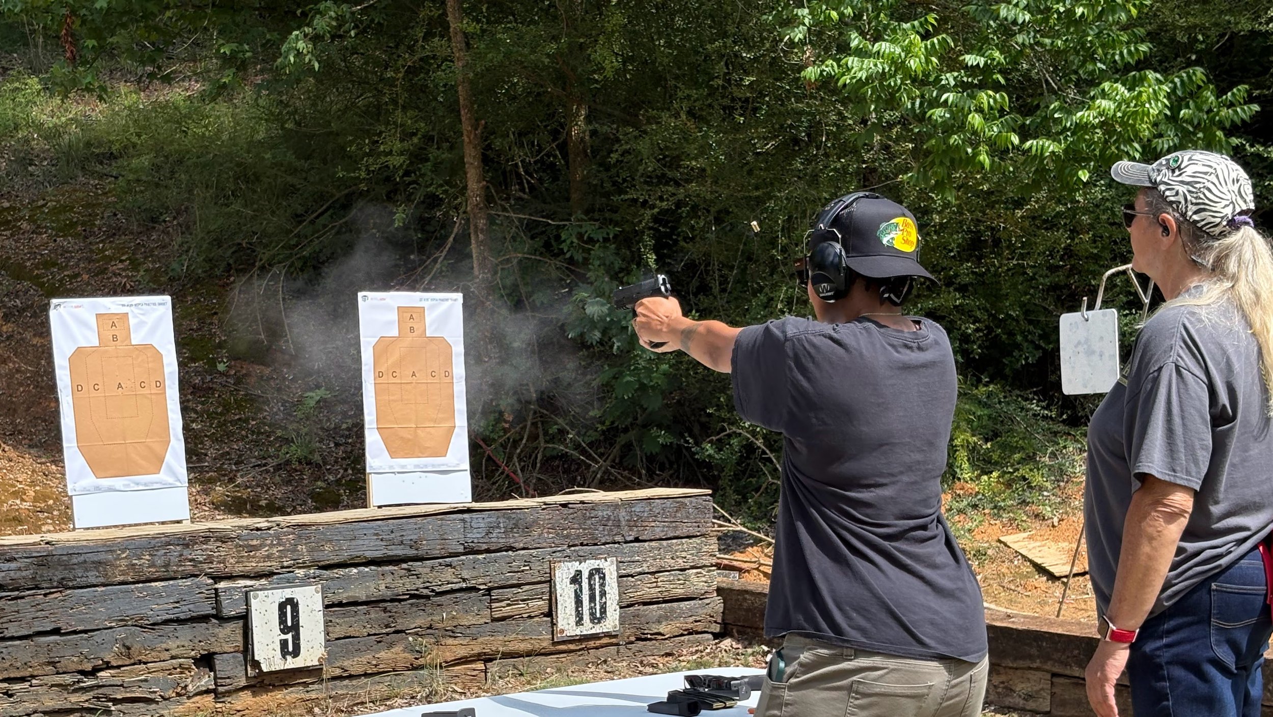 Two women at outdoor shooting range, one aiming a handgun, the other watching, with paper targets and numbered distance markers behind them.