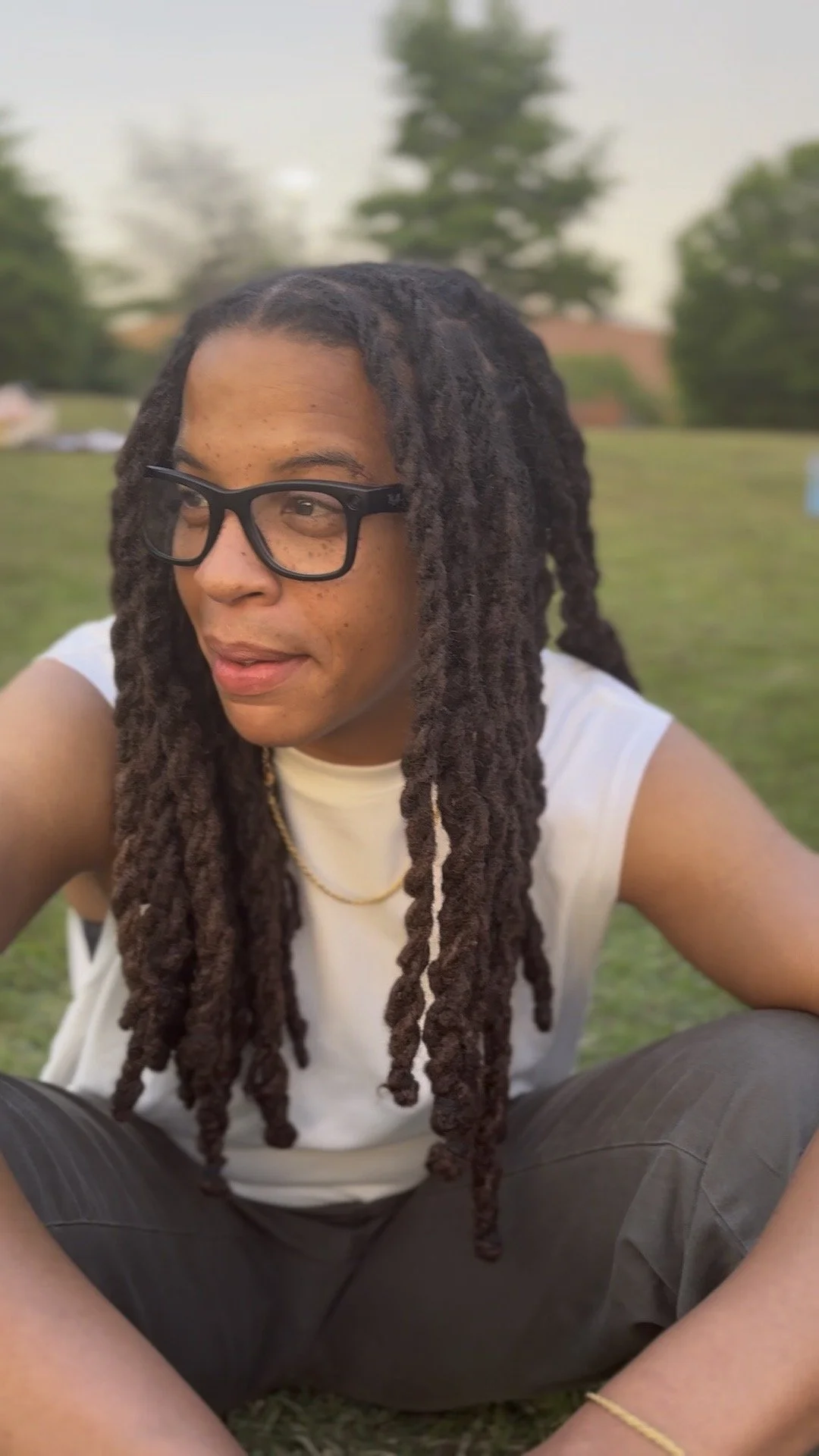 A person with medium skin, long dreadlocks, glasses, and a white sleeveless shirt sitting outdoors on a grassy field during sunset.