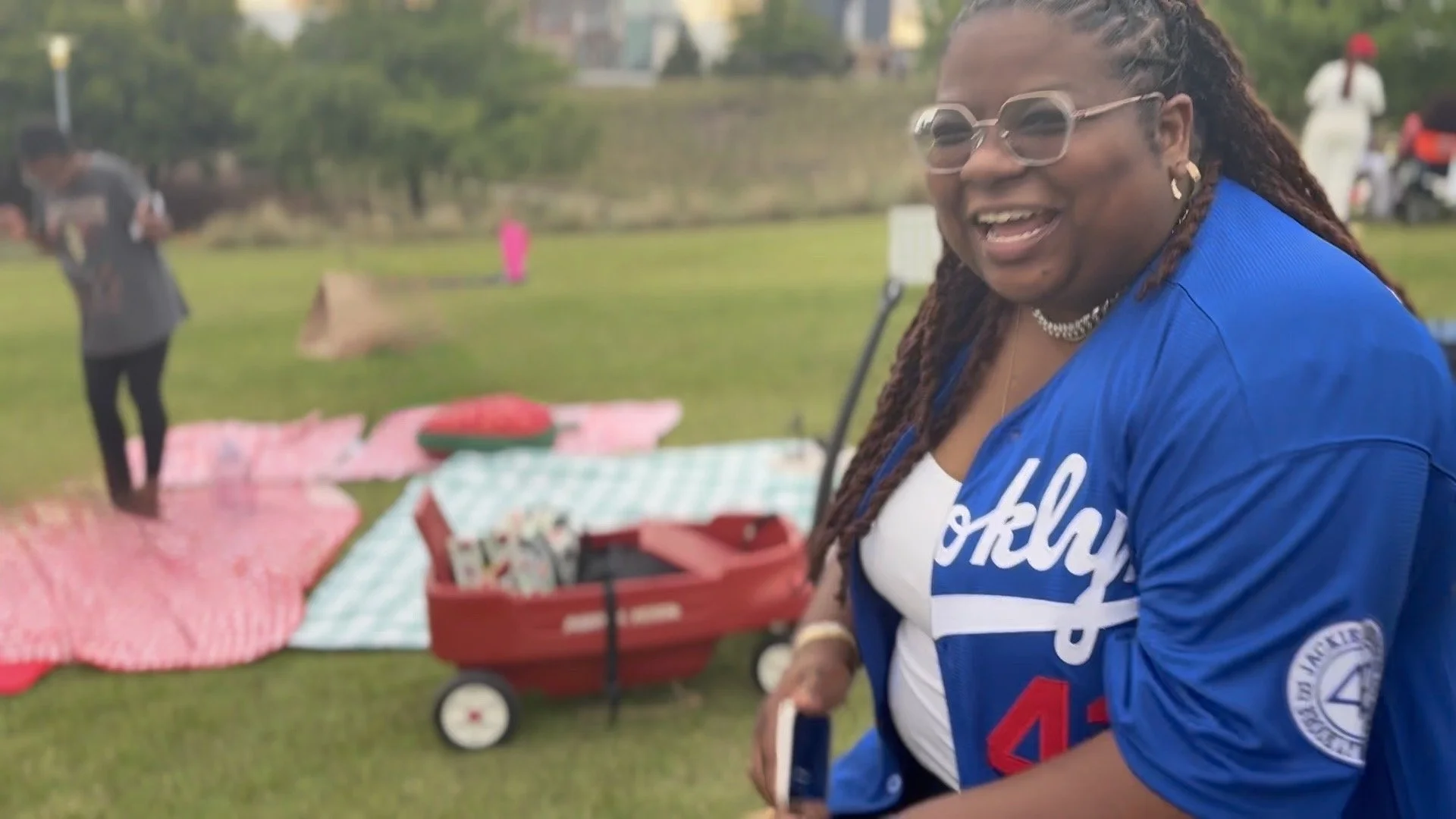 Smiling woman with glasses and braided hair, wearing a blue jacket with white text, at an outdoor event with picnic blankets and a red wagon in the background.