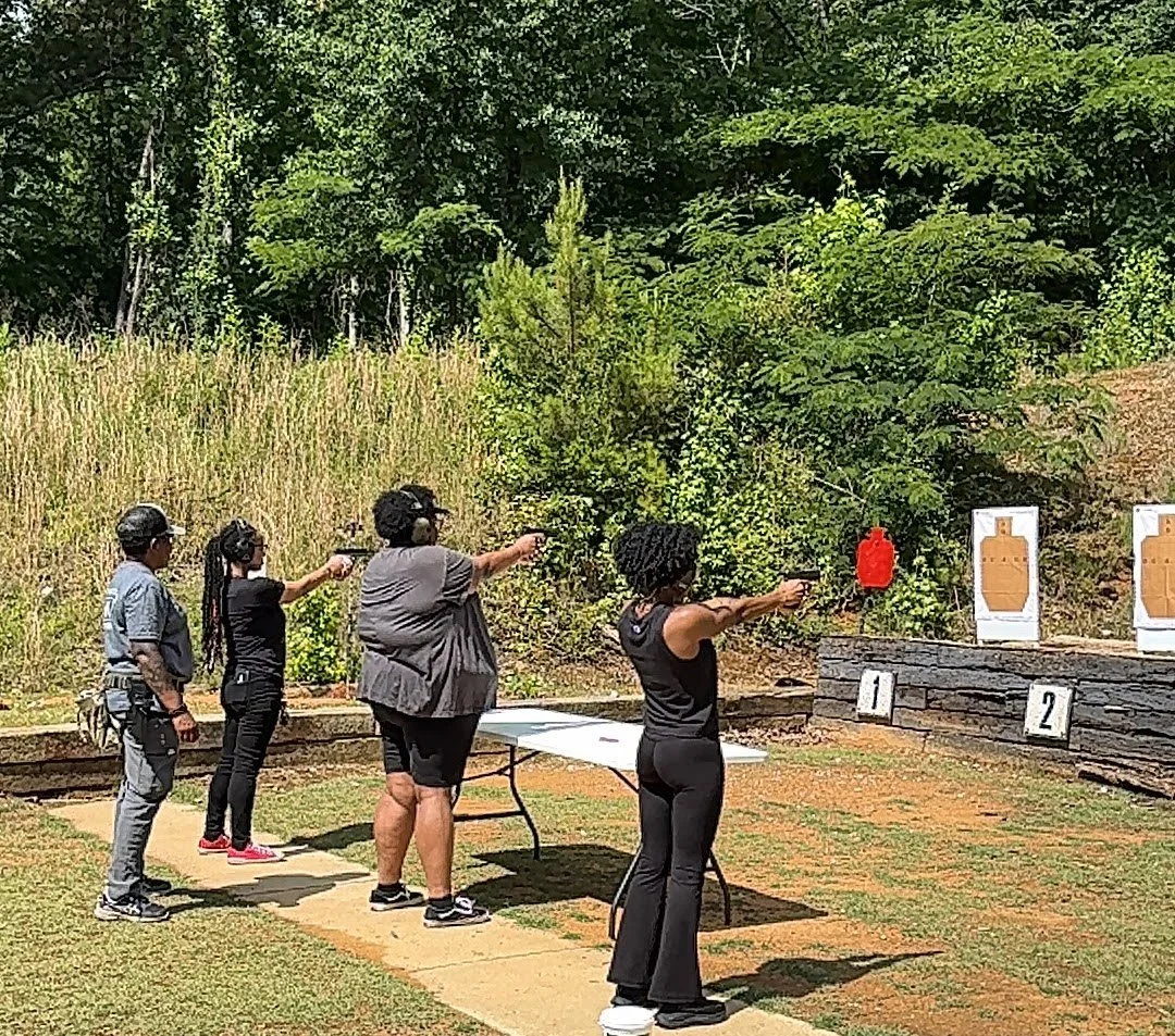 Women of The L Factor at an outdoor shooting range aiming at paper targets with guns.