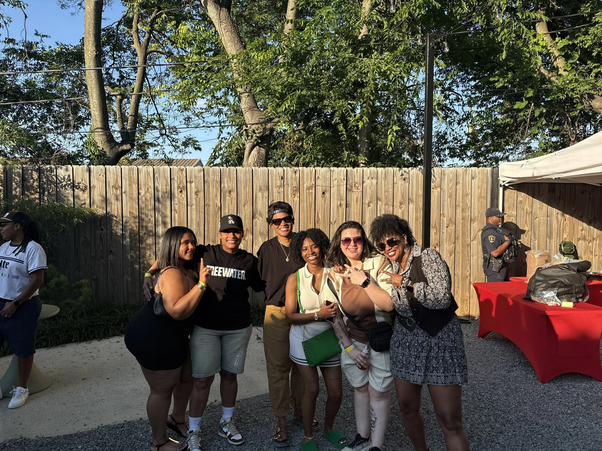 A group of seven diverse women standing outdoors behind a wooden fence, smiling and posing for a group photo at a social gathering, with trees and a clear sky in the background.