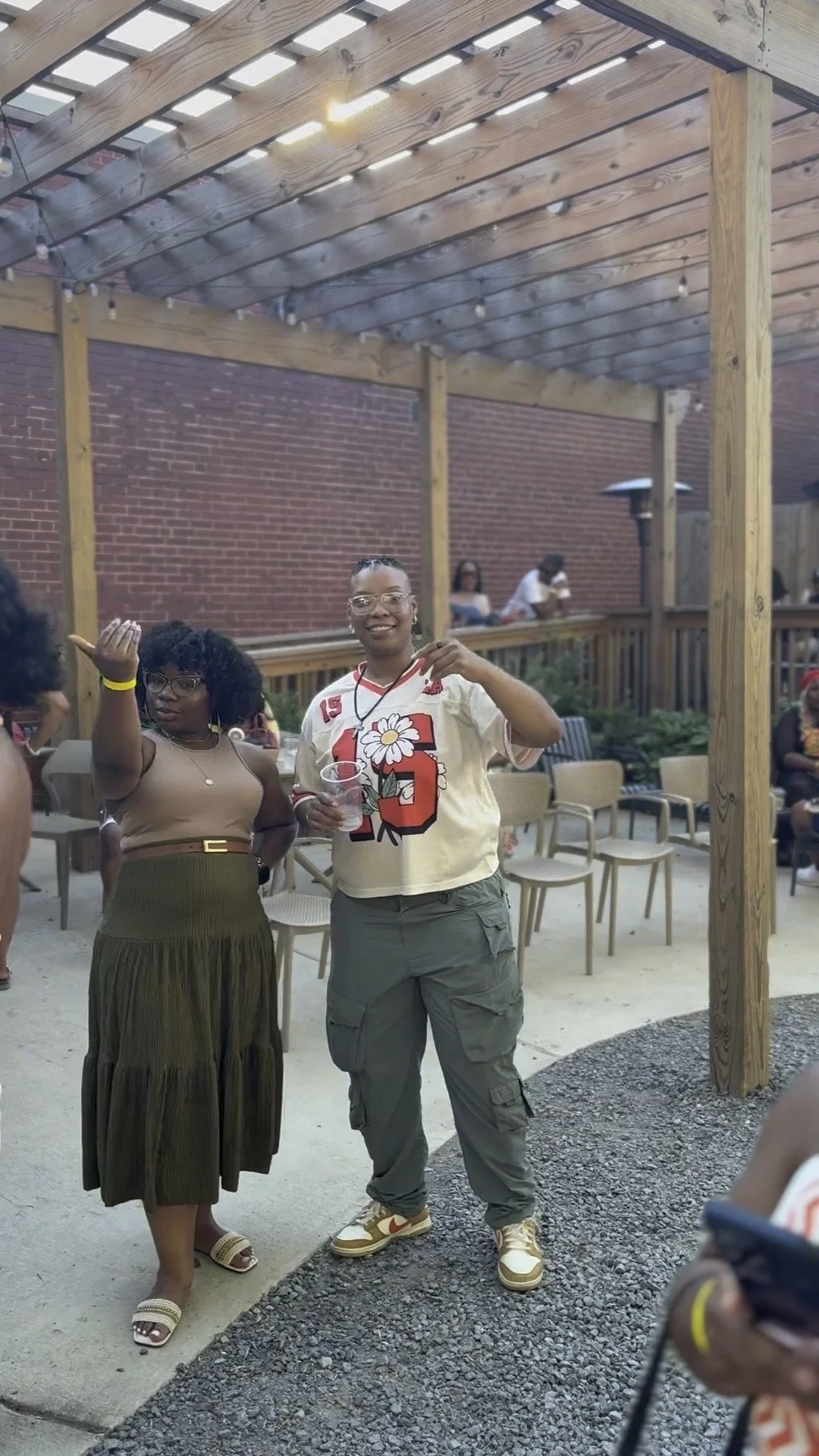 Two women standing outside under a wooden pergola at a social gathering, with chairs in the background and some people seated behind them.