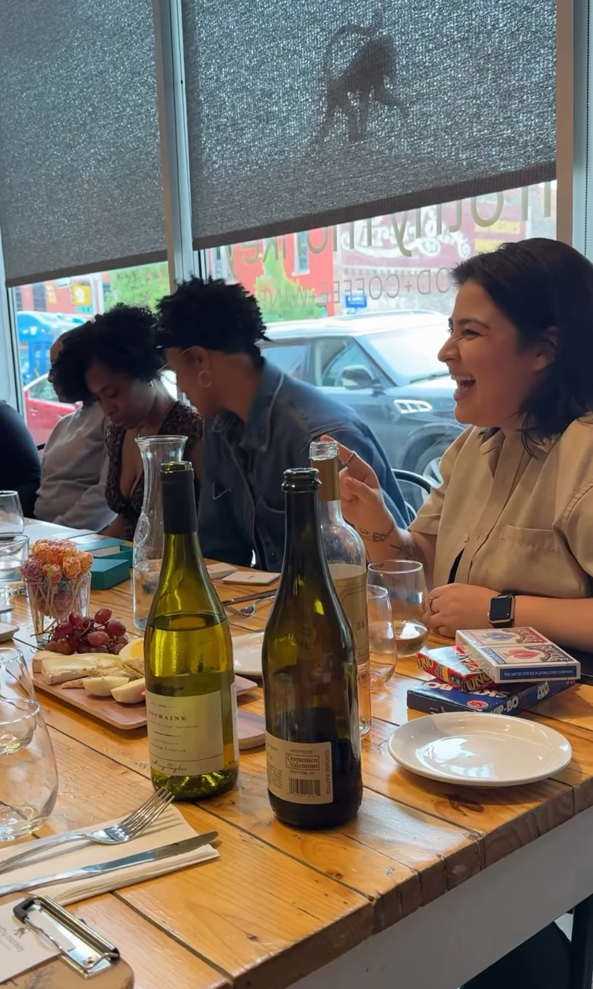People sitting at a wooden table with bottles of wine, grapes, cheese, and snacks in a restaurant. A woman on the right is smiling and laughing, with her drink in hand, while others are looking down or talking. Large windows with a view of parked car