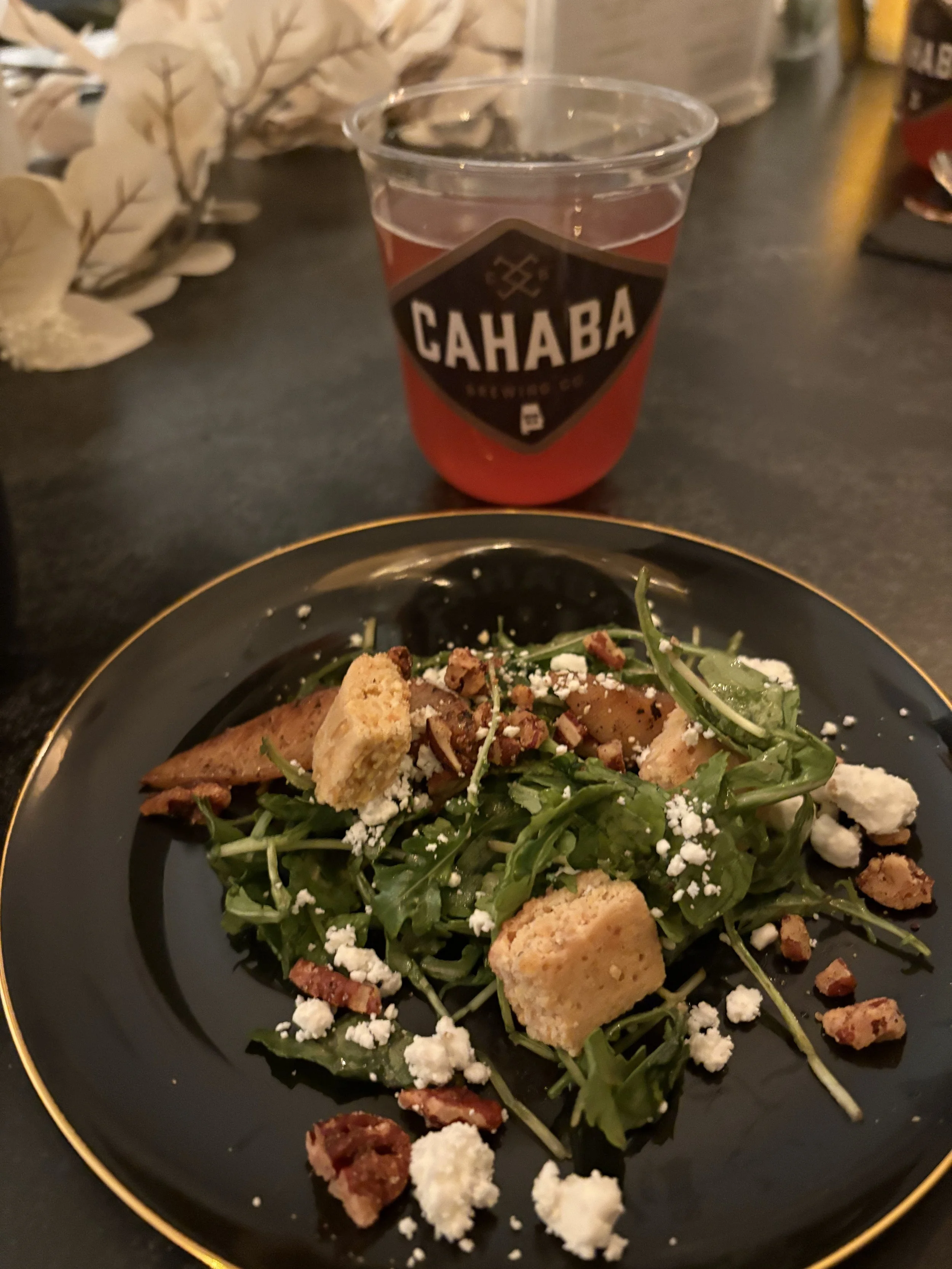 Plate of salad with arugula, crumbled cheese, bacon bits, and croutons, with a glass of pink beverage labeled 'Cahaba Brewing Co' in the background.