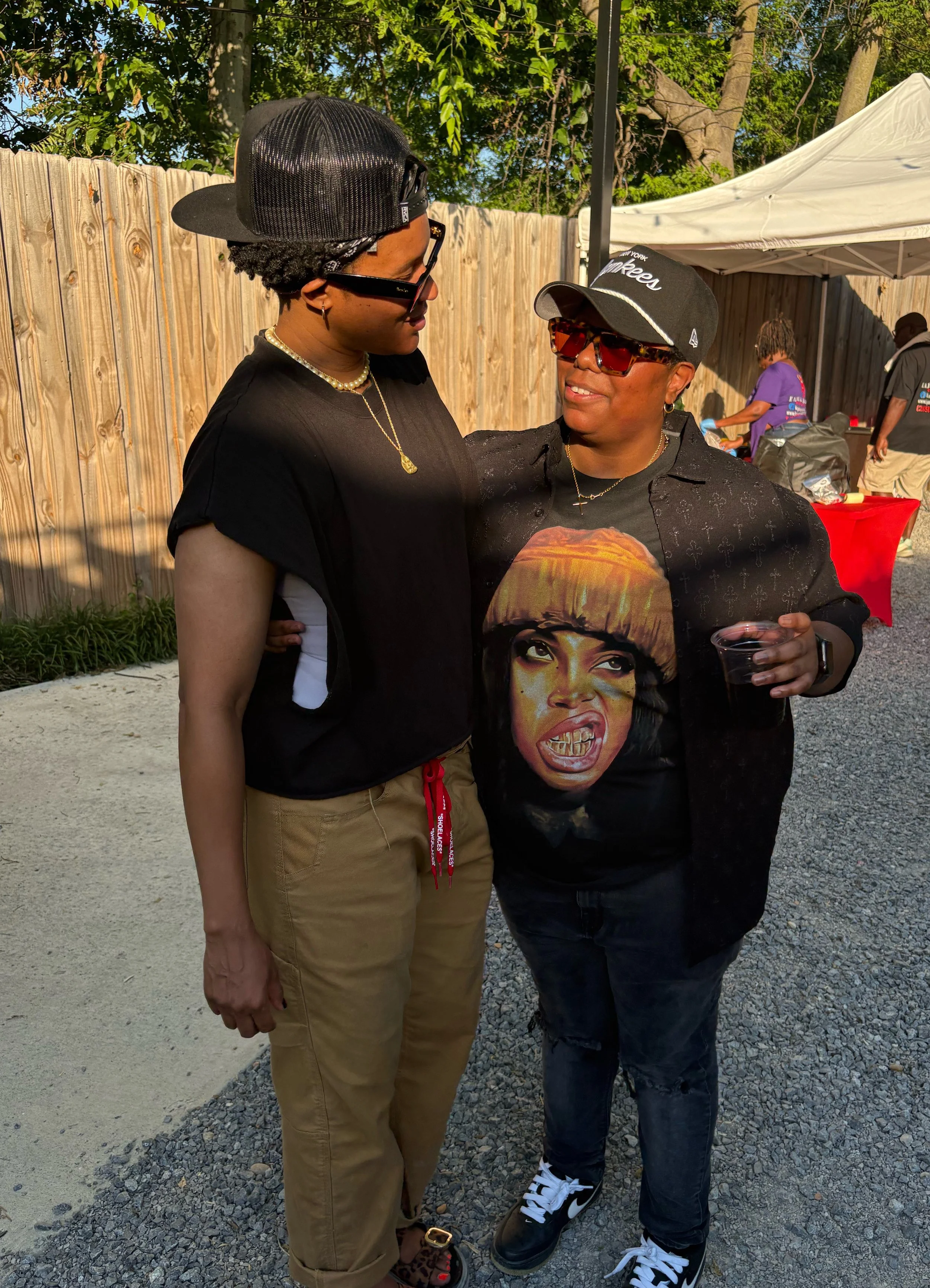 Two women facing each other, smiling, standing outdoors in front of a wooden fence. One woman is wearing a black cap, sunglasses, a graphic t-shirt, and has short hair and earrings. The other is wearing a black visor, glasses, a black Boutique shirt,