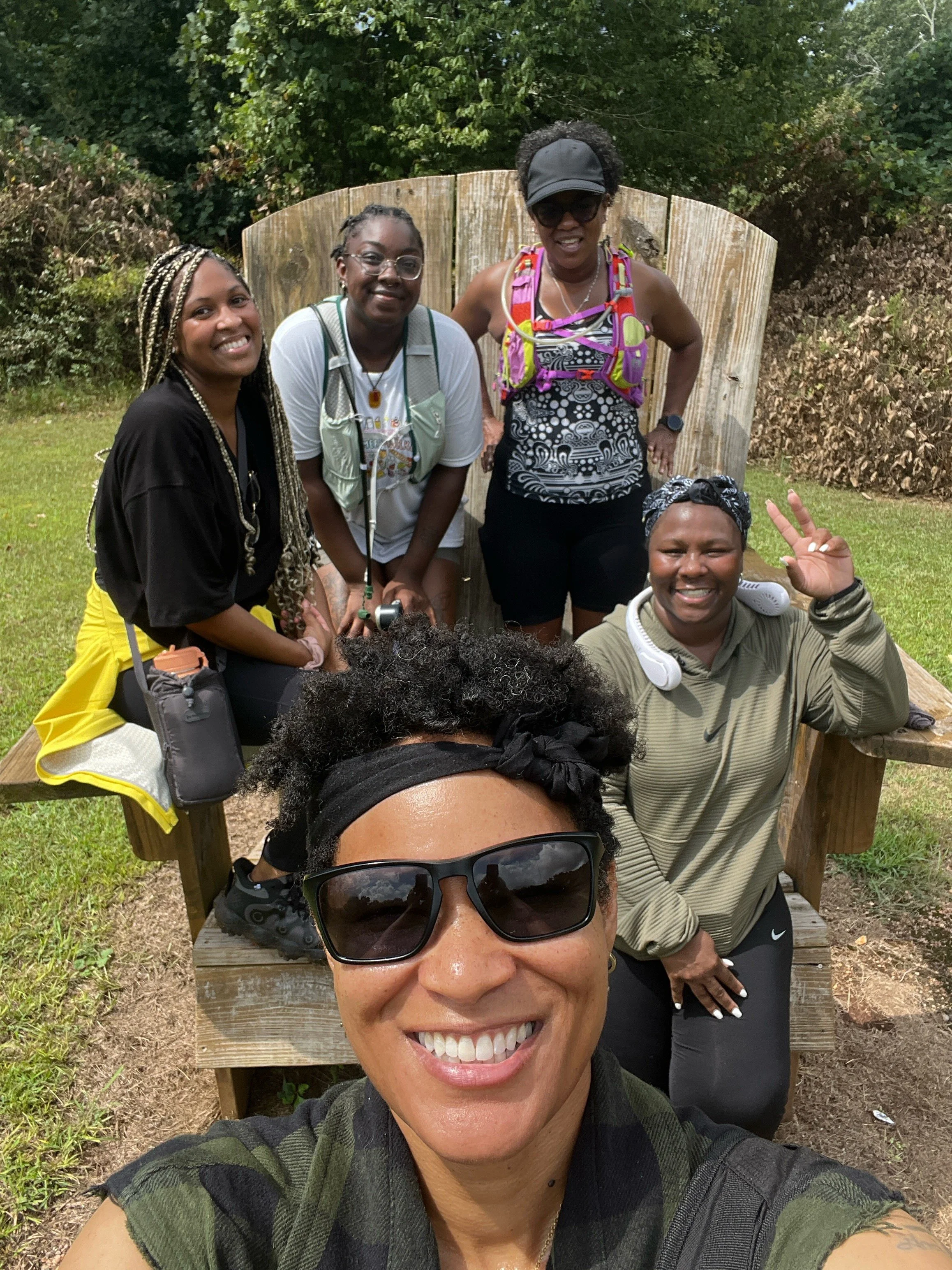 A group of five women outdoors, sitting on a wooden bench with a wooded background. One woman in front is taking a selfie, smiling with black sunglasses and a green camouflage shirt. The other women are smiling and wearing casual, colorful clothing a