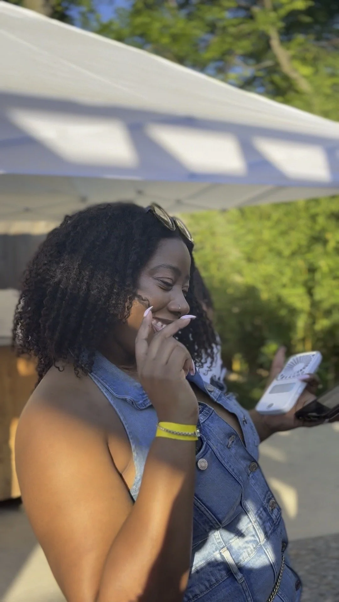 A woman with curly hair smiling, speaking on her phone, wearing a sleeveless denim vest and a yellow wristband, standing outdoors under a canopy during daytime.