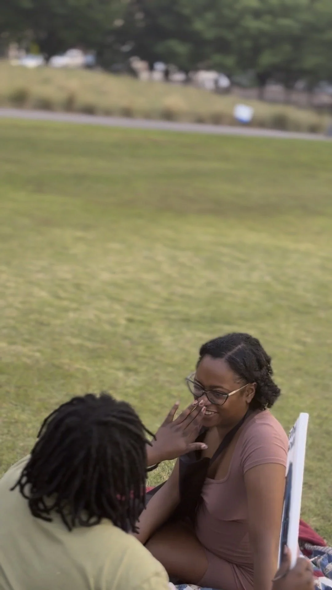 Two women sitting outdoors on grass, one with glasses and a light pink dress covering her mouth while smiling, and the other with dreadlocks and a yellow shirt, reaching towards the first woman's face, engaging in an affectionate moment.