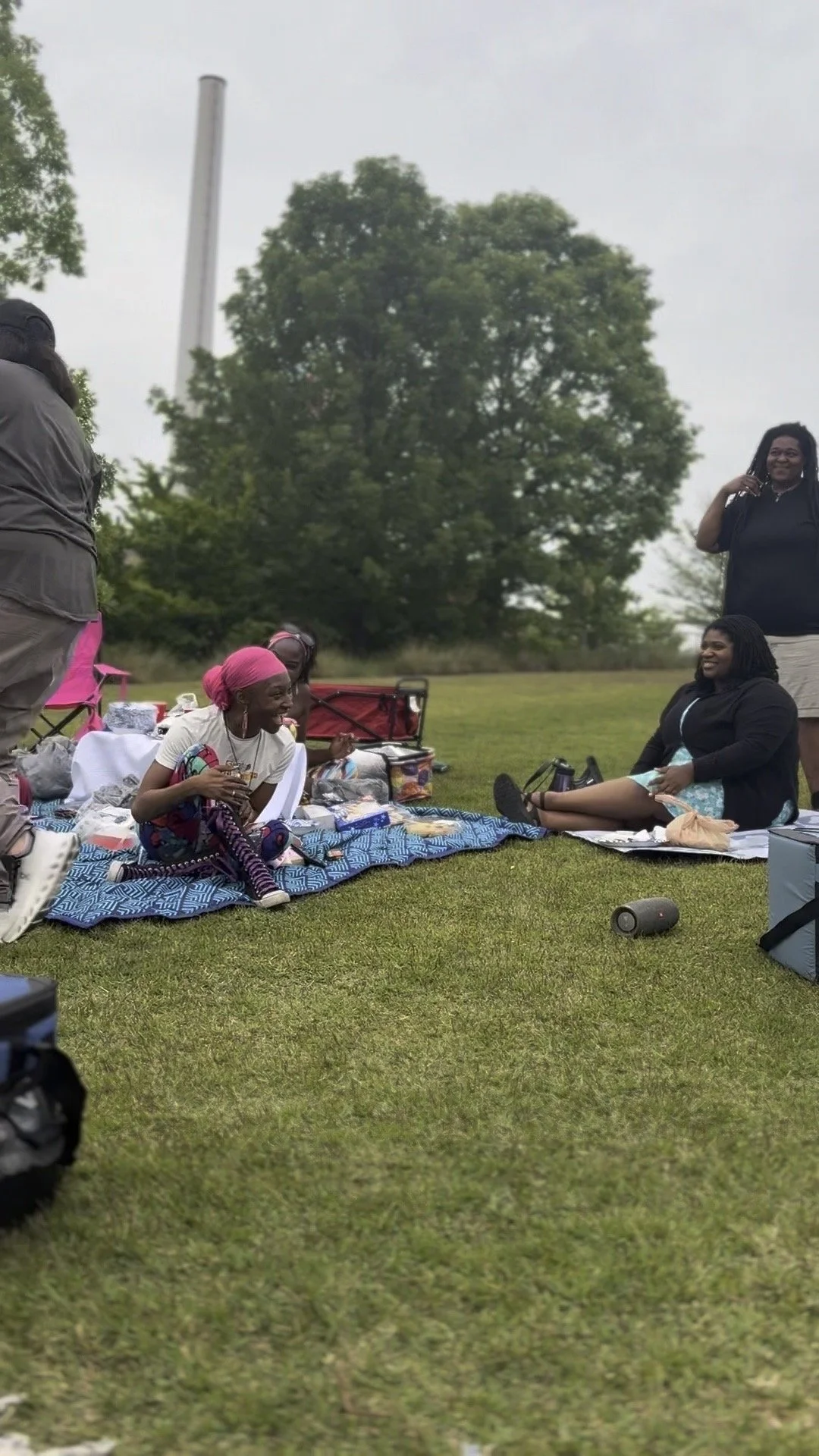 A group of women enjoying a picnic outdoors on a grassy field with trees in the background, sitting on a blanket and chatting, with food and drinks around them.