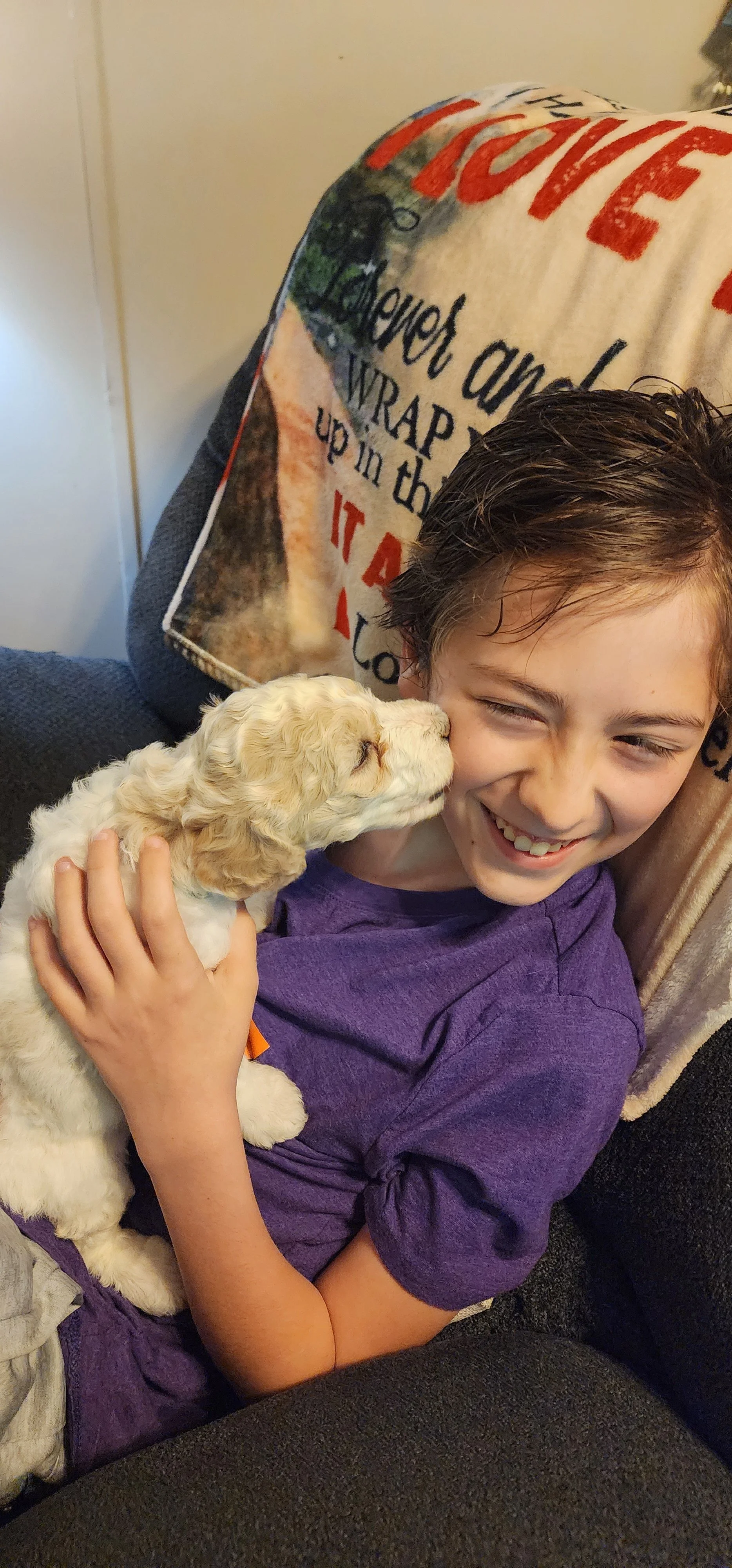 A boy in a purple shirt holding a small, light-colored puppy close to his face, both appearing to touch noses, sitting on a couch with a blanket draped over the backrest.