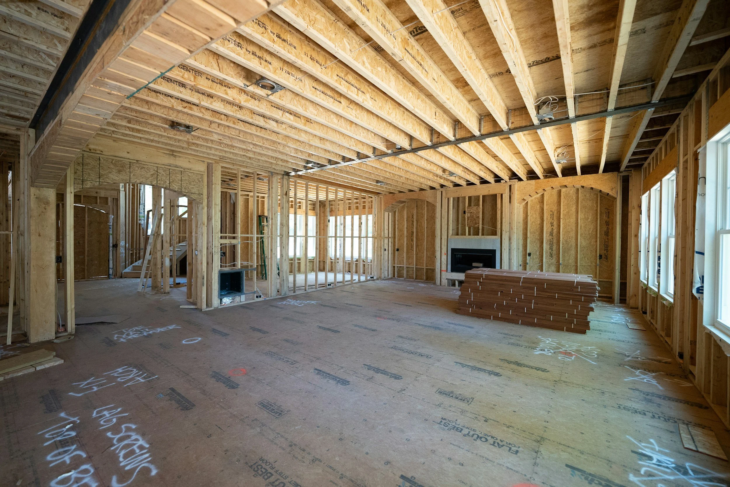 The interior of a house under construction with exposed wooden framing, including ceiling beams and wall studs, with some construction materials and a fireplace in the background.