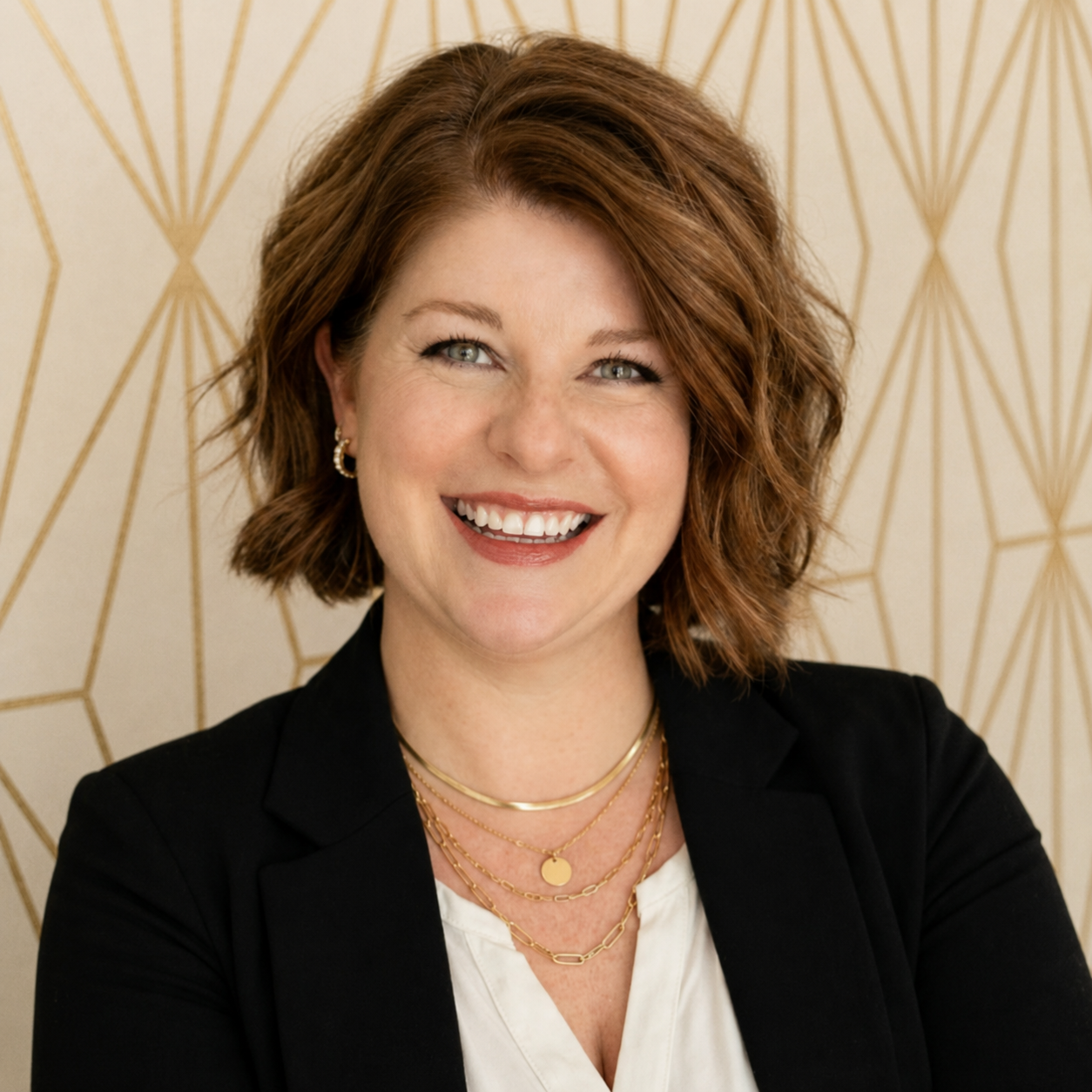 A smiling woman with shoulder-length, wavy, auburn hair, wearing a black blazer, layered gold necklaces, and small gold earrings, standing in front of a gold geometric patterned background.
