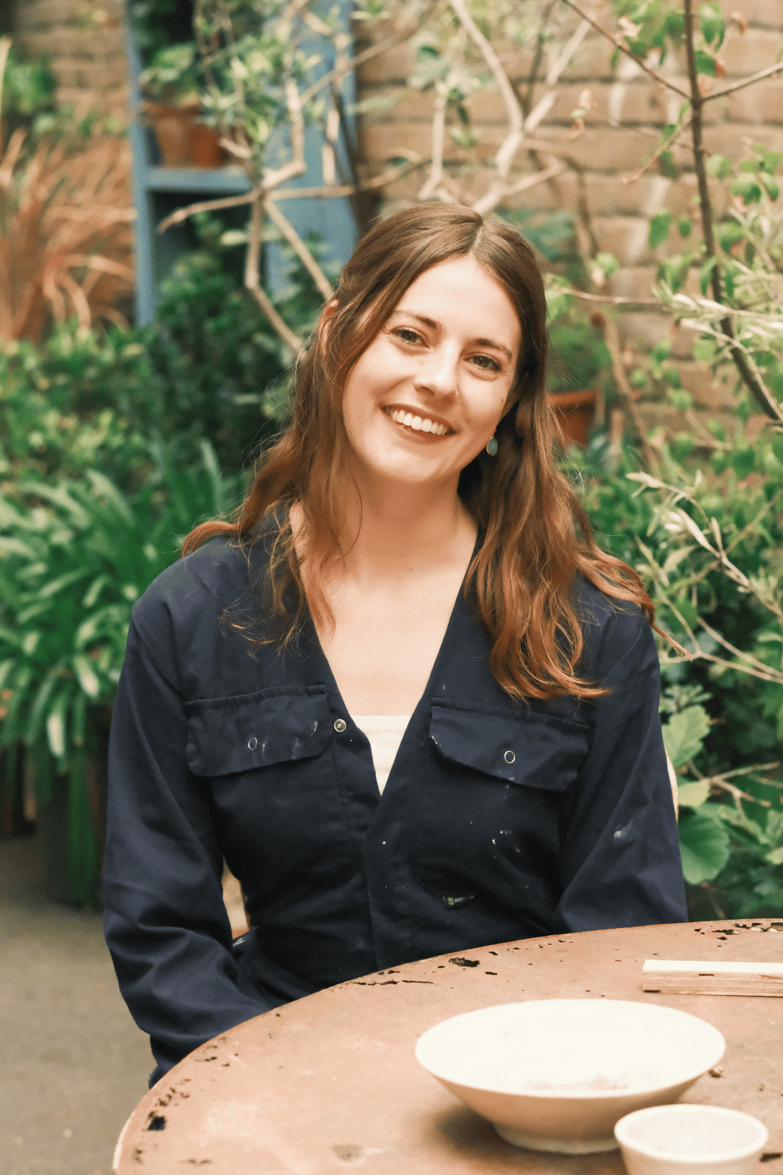 A woman with wavy brown hair and light skin, smiling and wearing a dark jacket, is sitting at a table in a garden or indoor plant setting. There is a white bowl and a smaller white bowl on the table in front of her.