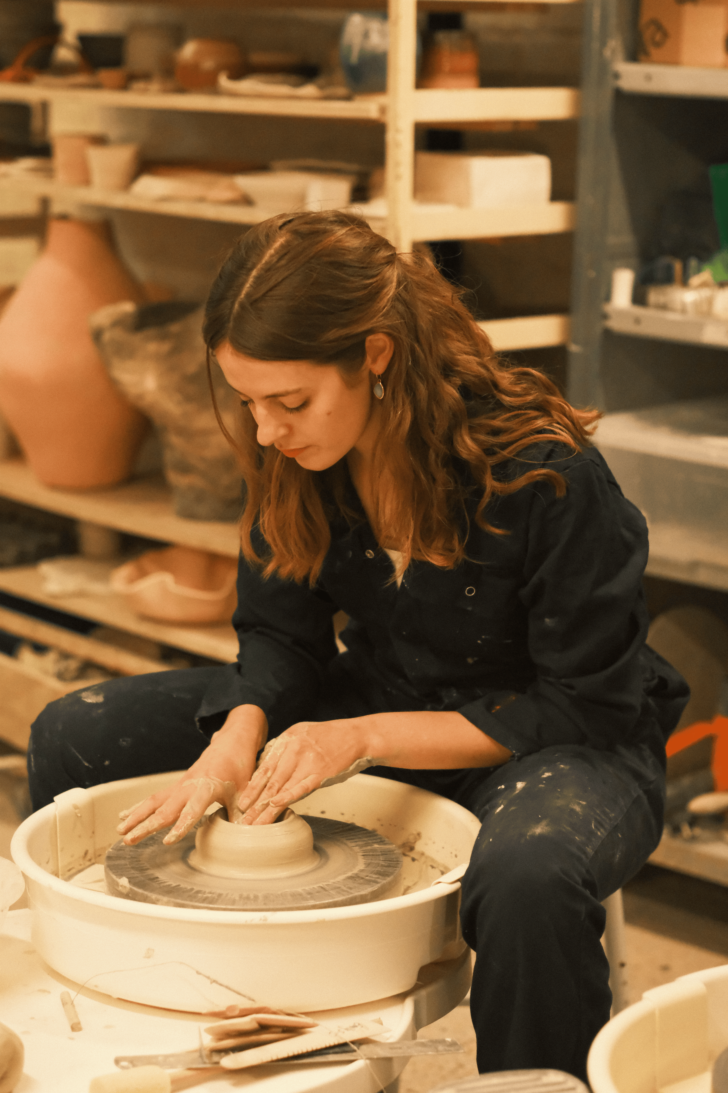 A woman with long, wavy brown hair, wearing a black shirt and black pants, is working on a pottery wheel shaping clay in a pottery studio. Shelves with various pottery pieces and tools are in the background.
