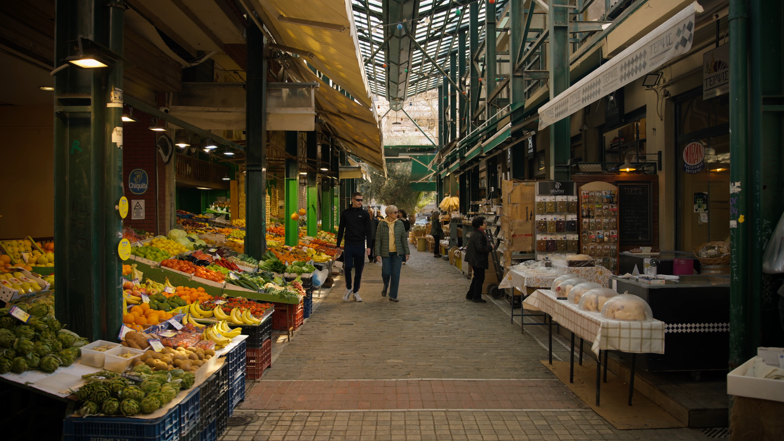 Outdoor marketplace with fruit, vegetables, and other goods on display, people shopping and walking under a canopy with string lights.