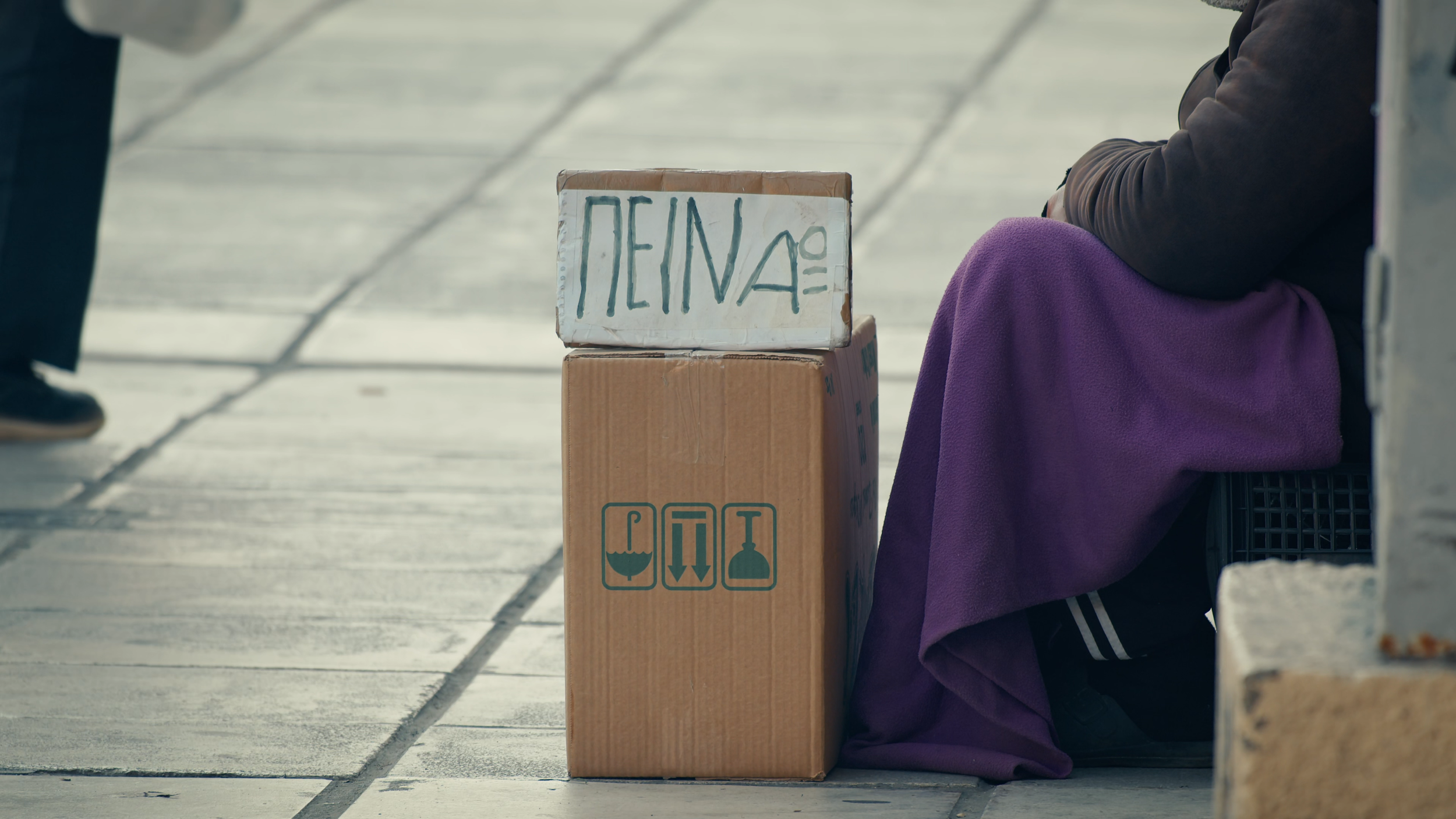 A person sitting on the ground with their knees up, wearing a purple blanket. Next to them are two cardboard boxes stacked, one with a white sign reading 'HOME' written backwards and symbols indicating fragile contents and upright position.