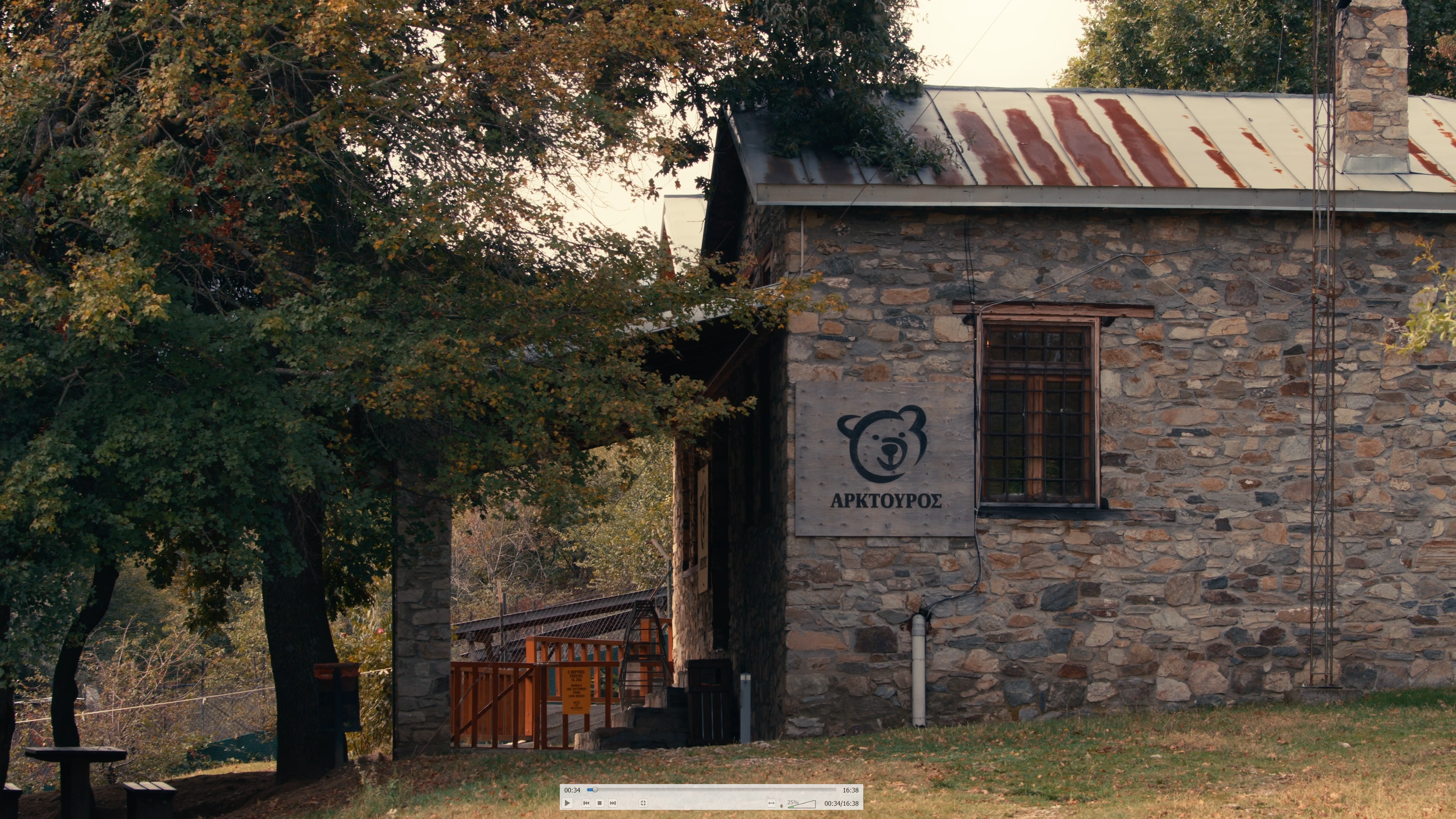 A rustic stone building with a corrugated metal roof, surrounded by trees with autumn leaves. There is a sign with a bear logo and Greek text on the wall.