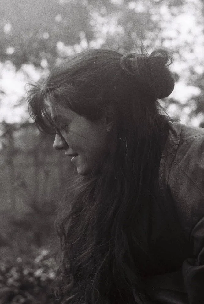 A black and white photo of a woman with long dark hair, smiling and looking downward outdoors, with blurred trees in the background.
