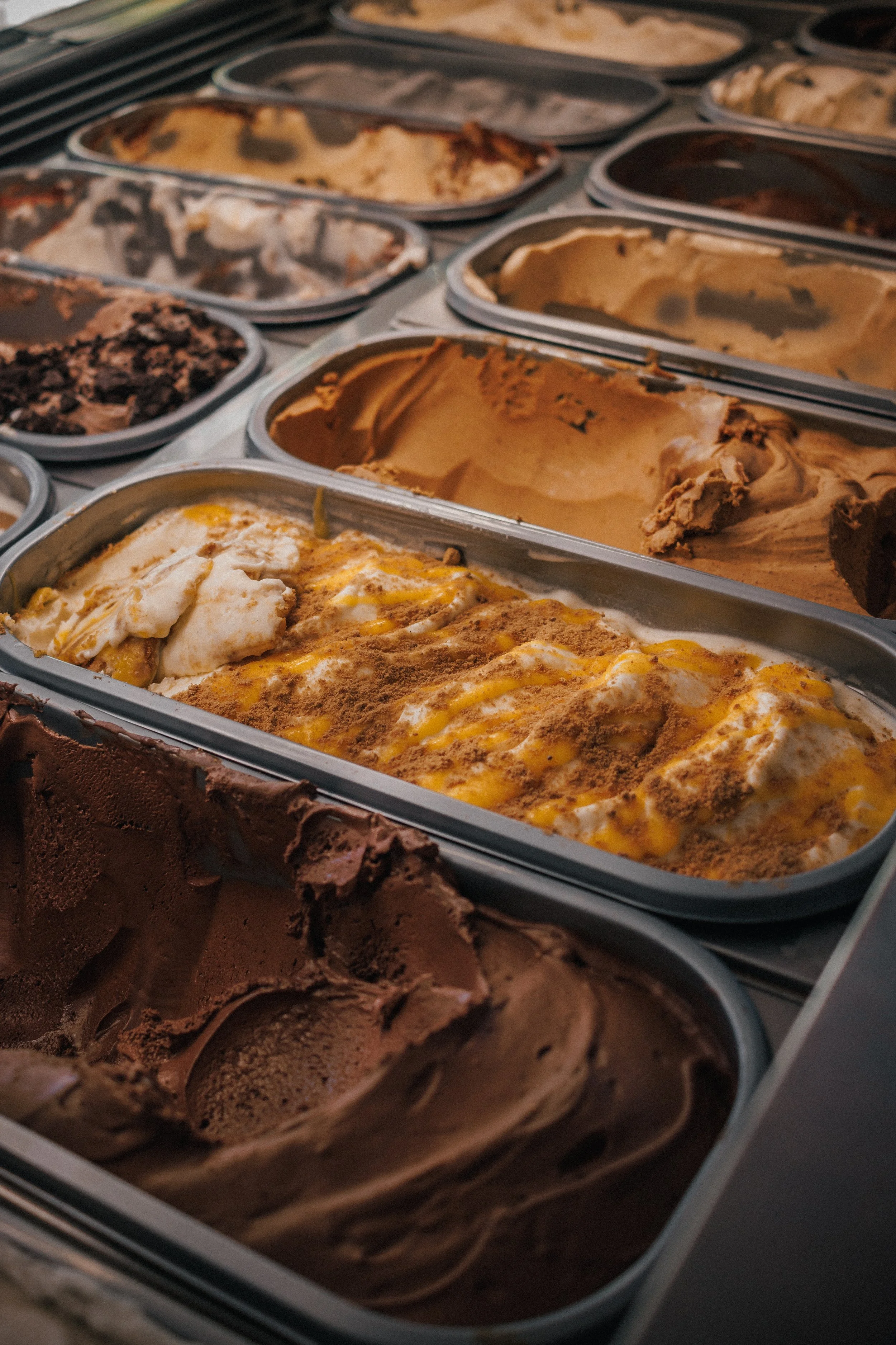 Multiple open containers of various flavors of ice cream, including chocolate, vanilla, and mixed flavors, displayed in a freezer.