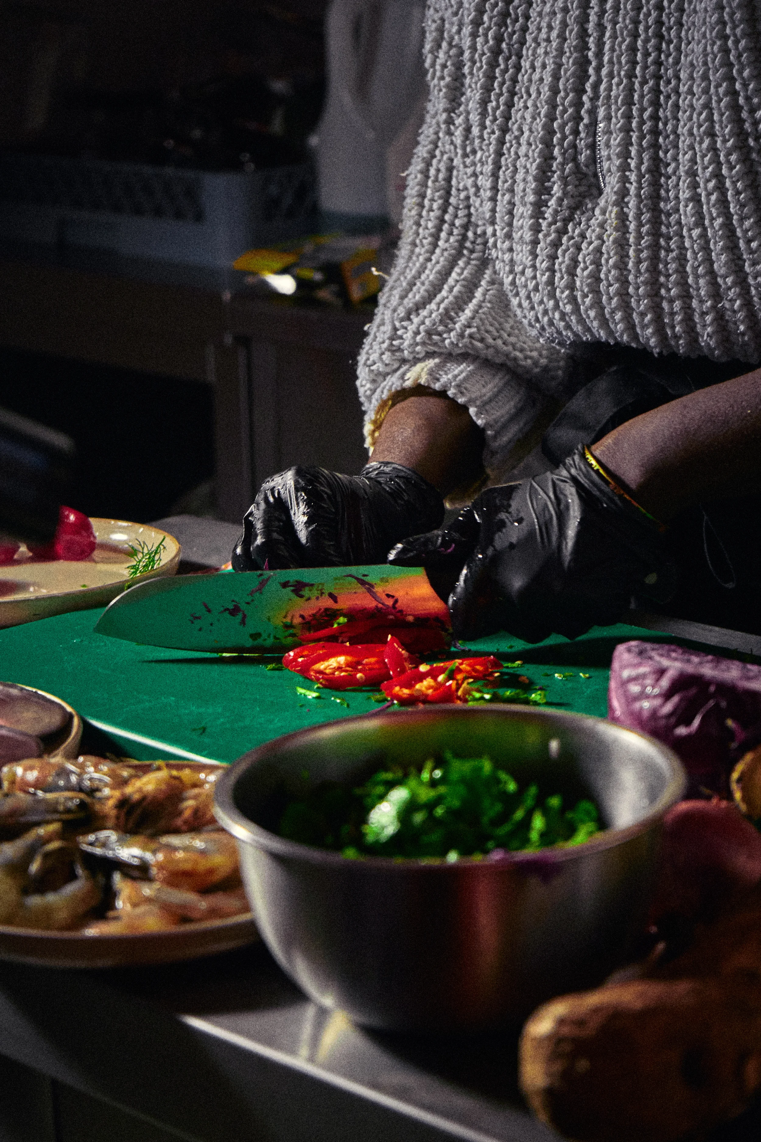 A person wearing a white sweater and black gloves is preparing food at a kitchen counter with various bowls of ingredients, including sliced red peppers and green herbs.